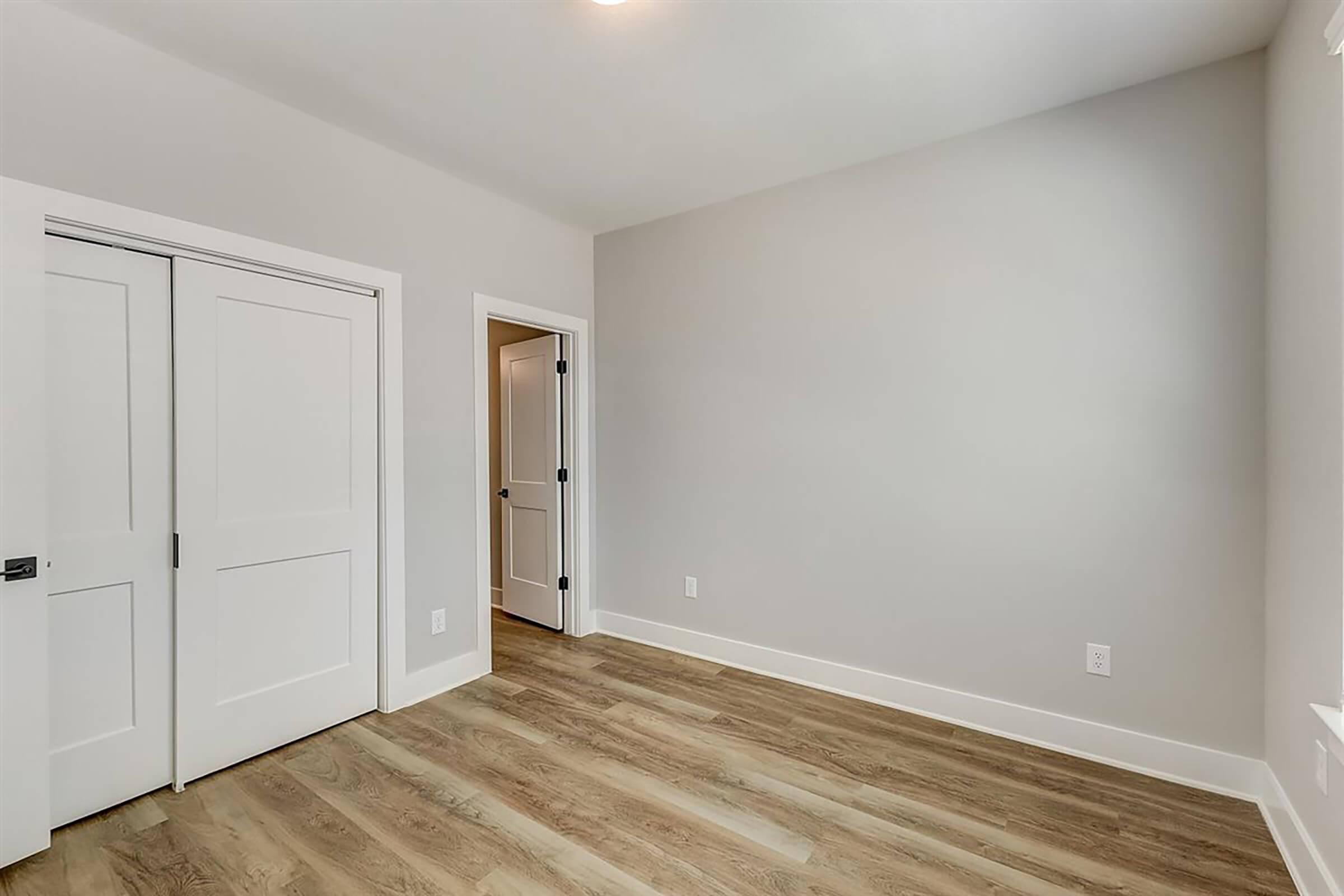 Empty room with light gray walls and laminate flooring. A closet with double doors is on the left, and a doorway leads to another room on the right. The space is well-lit, with a soft ceiling light, creating a clean and modern atmosphere.