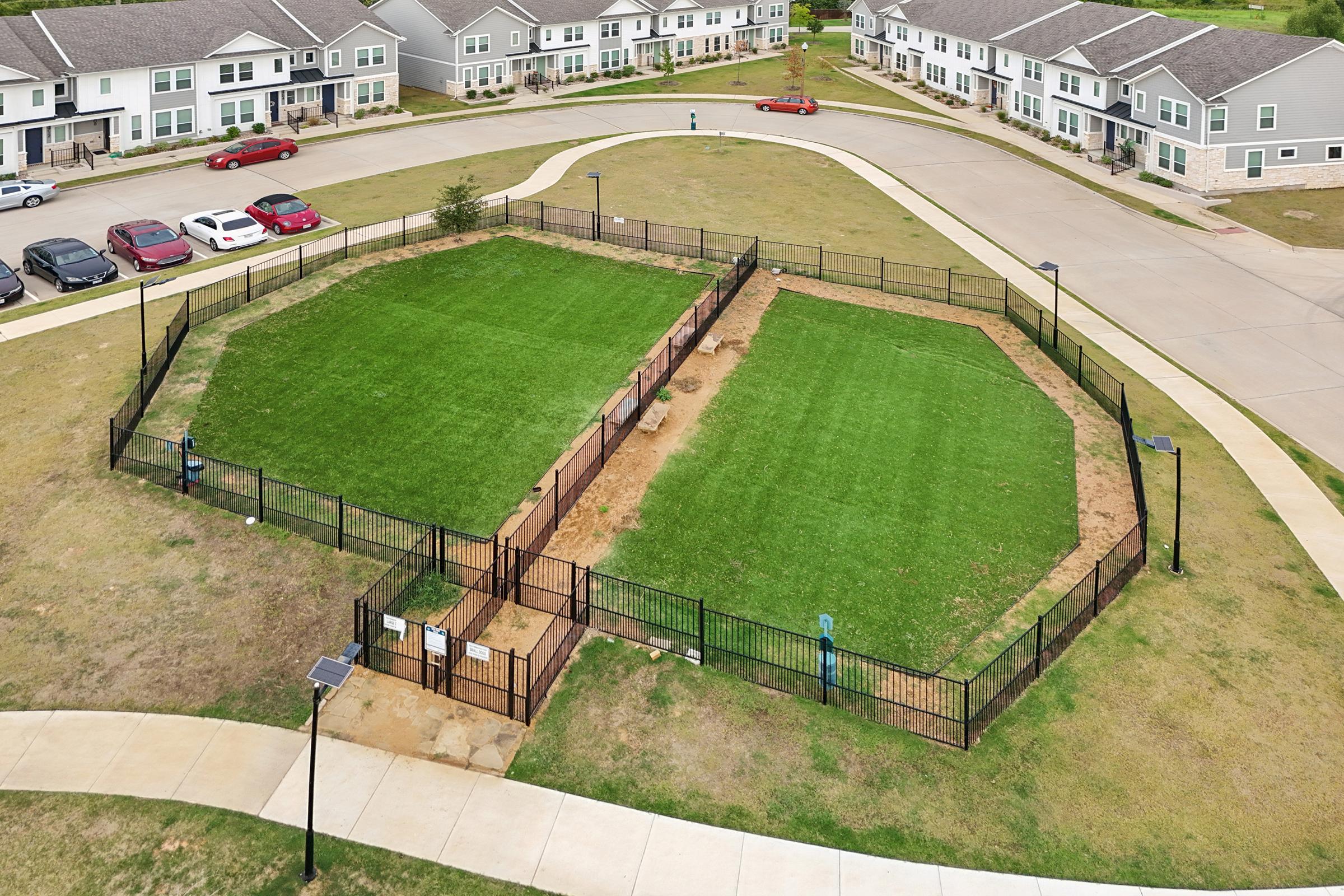 Aerial view of two fenced dog parks with green grass, surrounded by walking paths and residential buildings in the background. The left park has some visible equipment, while the right park appears more spacious. There are several parked cars nearby, and the overall area is well-maintained.