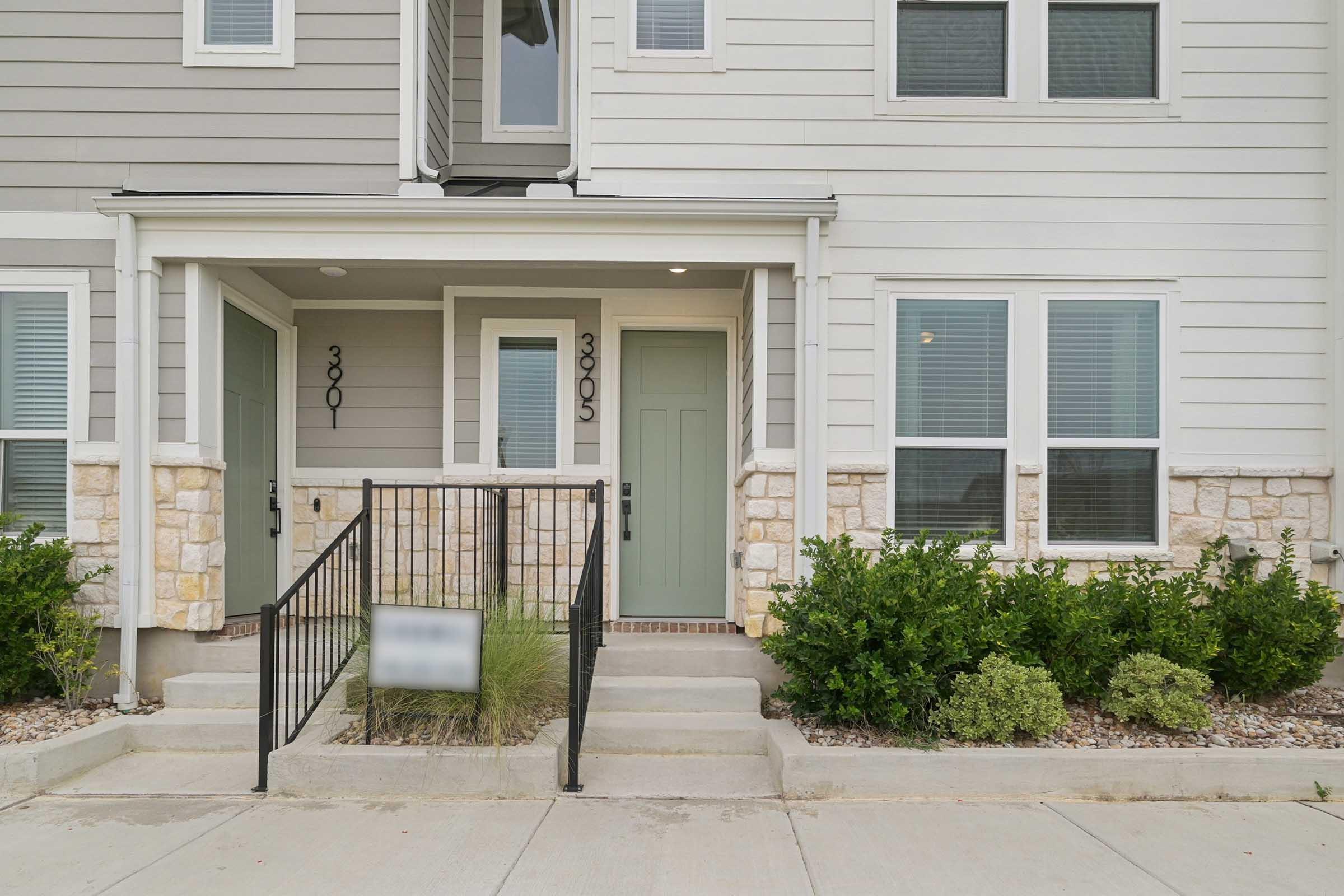 Front entrance of a two-story townhouse with light gray siding, featuring two doors marked 3001 and 3005. There are small shrubs and landscaping elements along the walkway and near the entrance. A black metal railing leads up to the steps, and the area is well-maintained with a clean concrete pathway.
