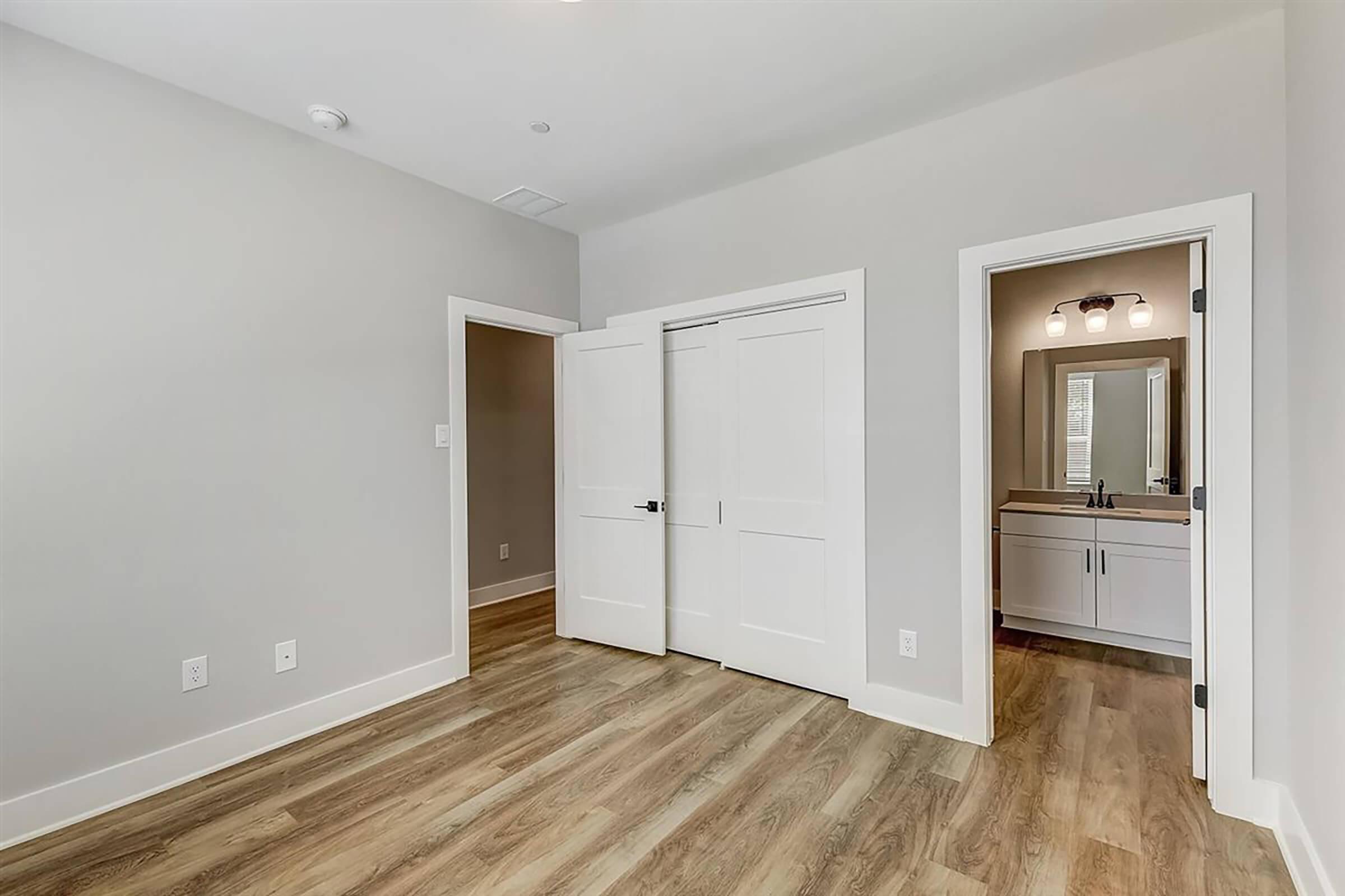 Empty room with light gray walls and a wooden floor. Two white doors are slightly open, leading to another room and a bathroom area with a double sink and mirror. Natural light fills the space, creating a bright and spacious atmosphere.