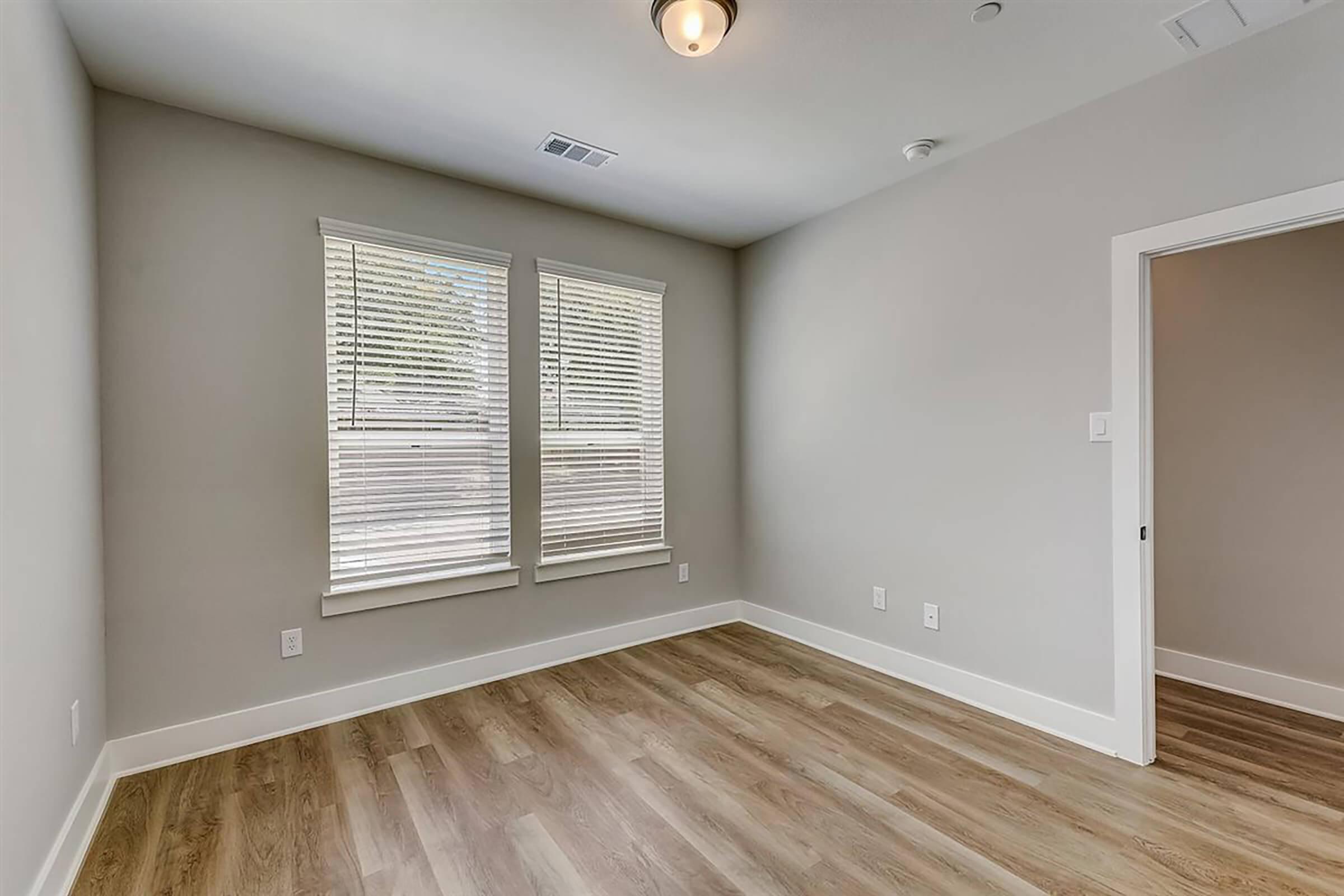 Empty room with light gray walls, two windows with horizontal blinds allowing natural light, and a wooden floor. Features a ceiling light fixture and a door leading to another space, creating a bright and spacious atmosphere.