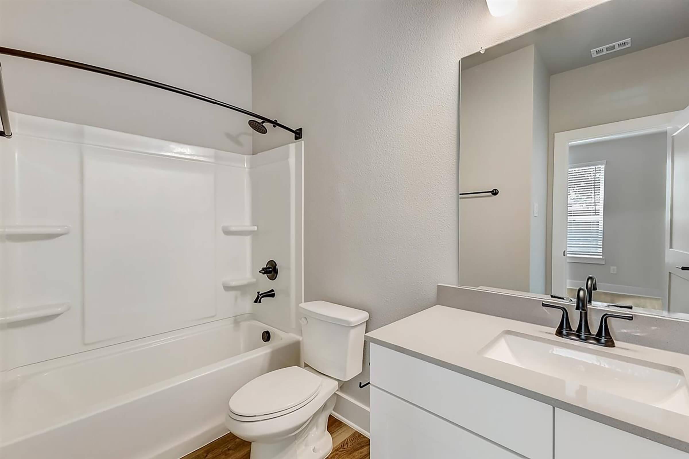 A clean, modern bathroom featuring a white bathtub with a shower, a toilet, a sleek vanity with a sink, and a large mirror. The walls are painted light gray, and the flooring is a warm wood tone. Natural light comes in from a window in the background, creating a bright and inviting atmosphere.