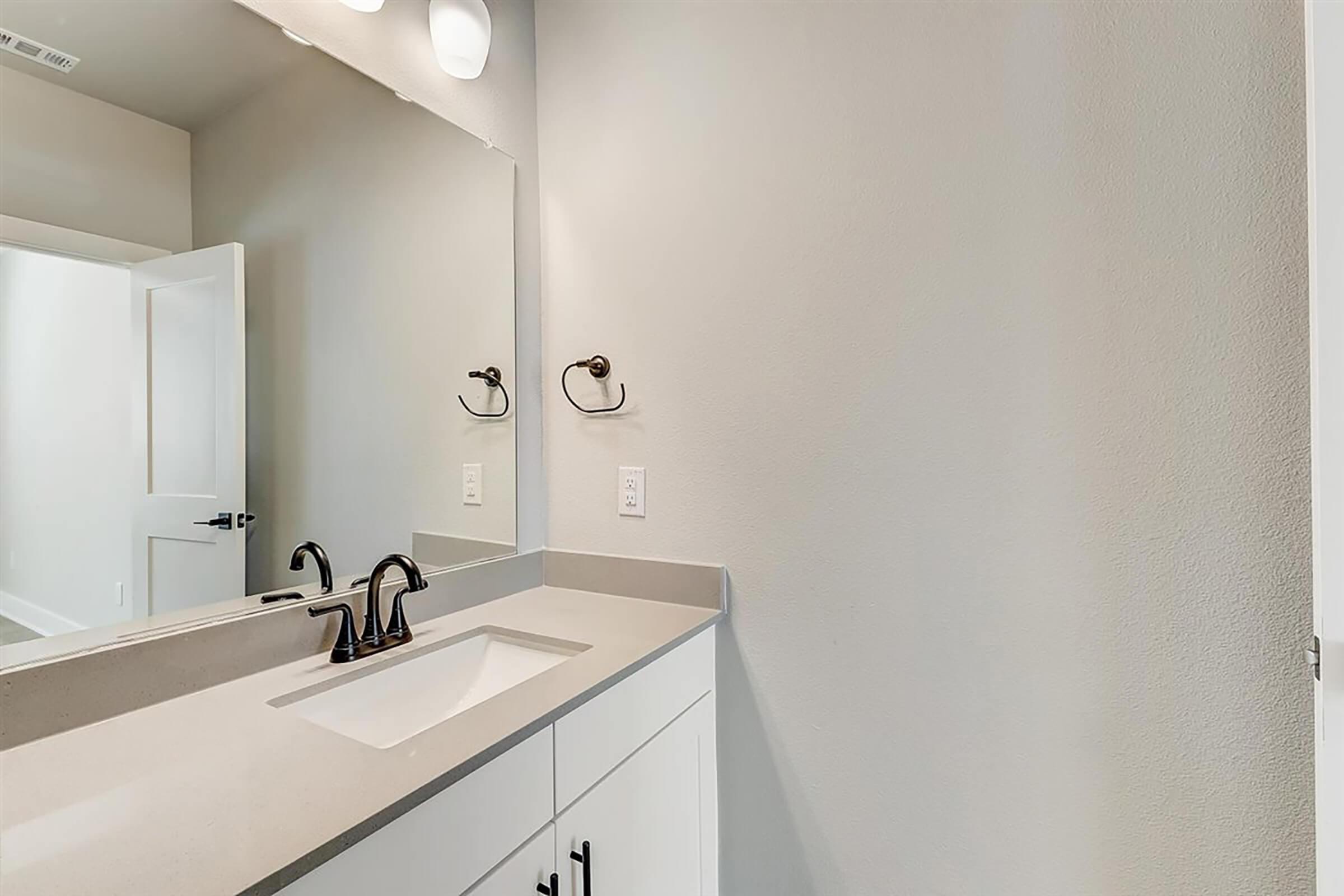 A modern bathroom featuring a light-colored wall, a sleek countertop with a rectangular sink, and a large mirror. The bathroom has minimalist fixtures and a simple design, with a towel holder on the wall and a doorway leading to another room. The overall aesthetic is clean and contemporary.