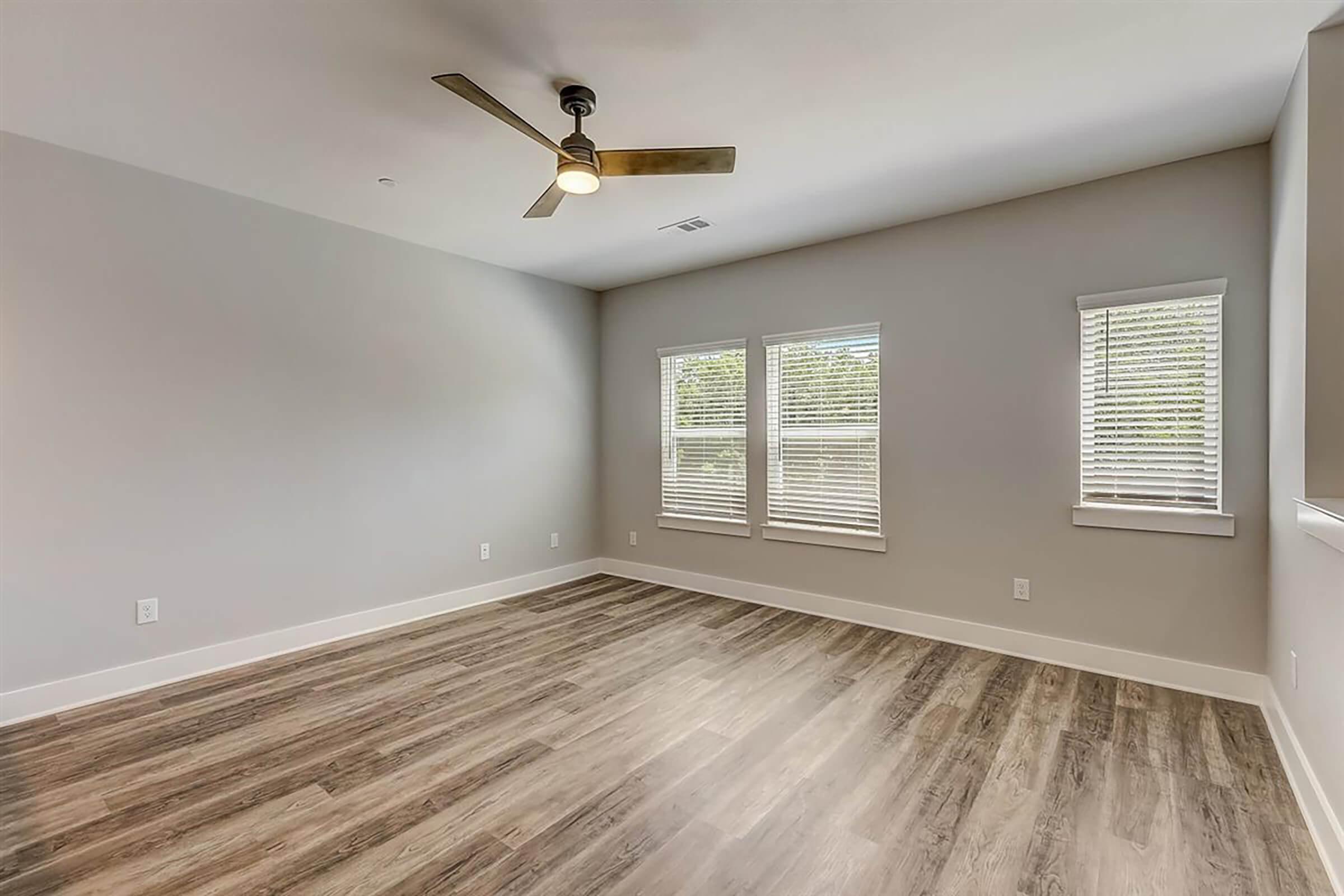 A spacious, empty room with light-colored walls, hardwood flooring, and a ceiling fan. It features three windows with white blinds allowing natural light. The room has a modern and minimalist design, making it suitable for various purposes, such as a living area or office.