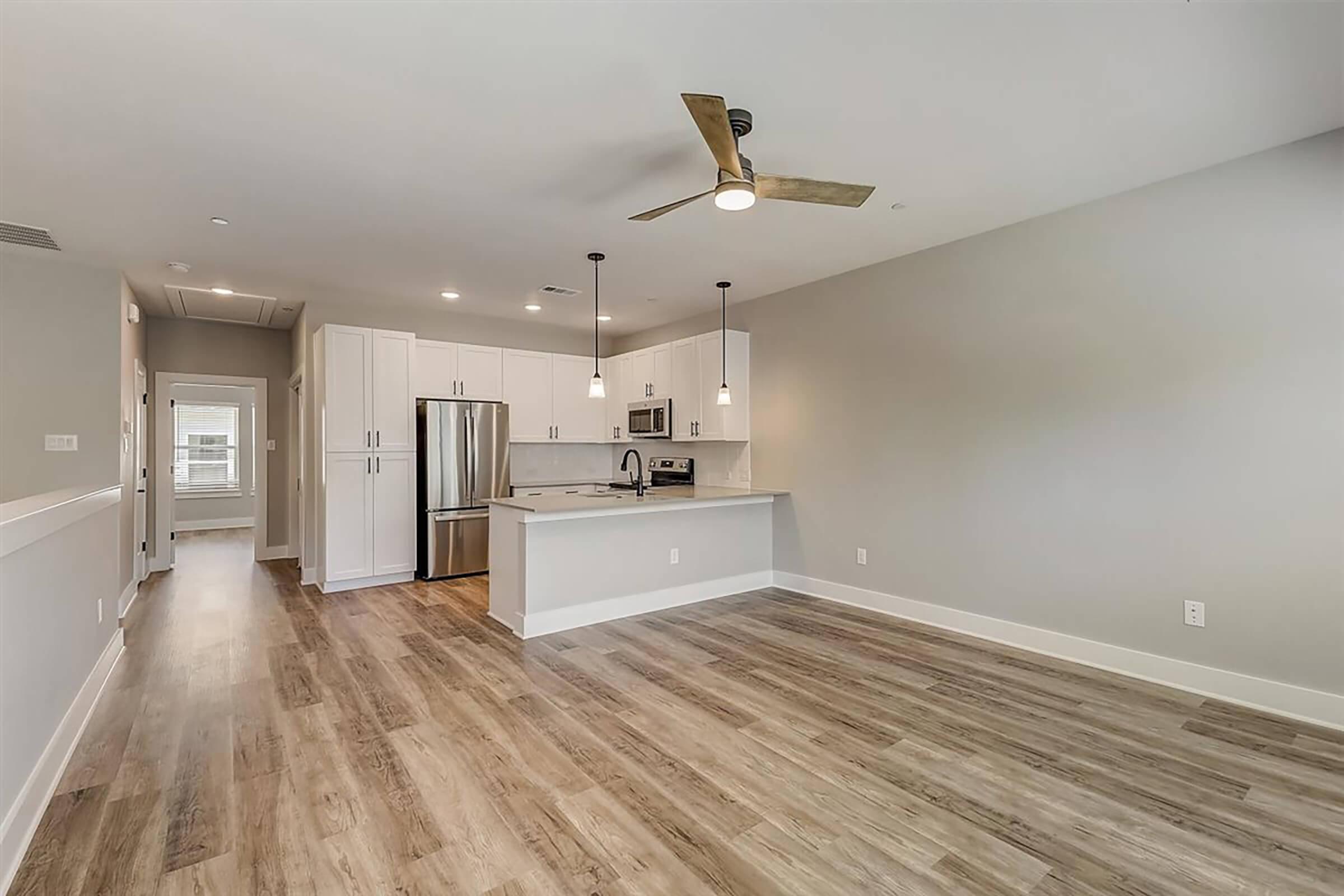 A modern, open-concept kitchen and living area featuring a ceiling fan, light wood flooring, and neutral-colored walls. The kitchen has white cabinets, stainless steel appliances, and a countertop with seating. Natural light streams in through a window, enhancing the spacious feel of the room.