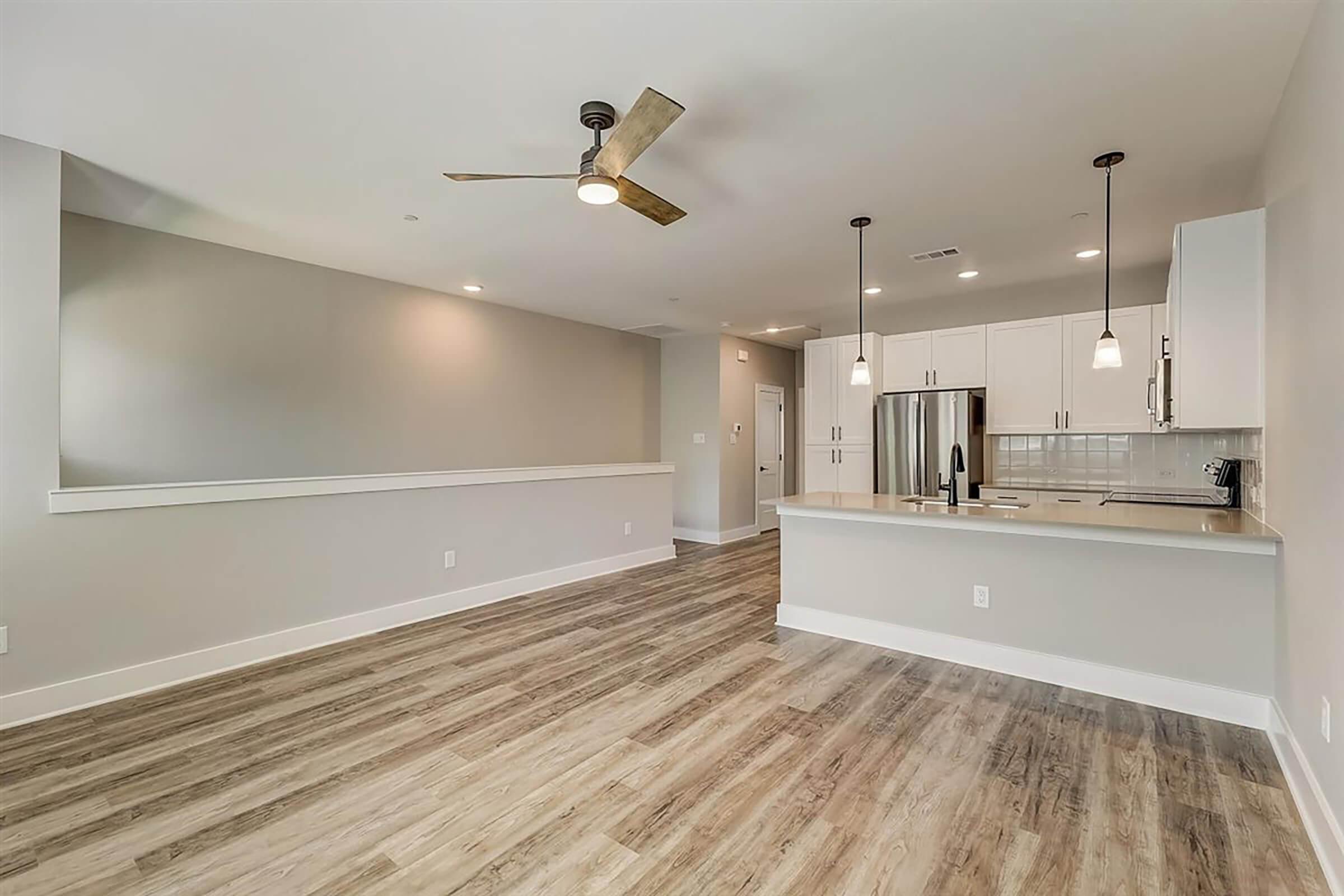 Modern interior of a spacious living area featuring a light-colored kitchen with stainless steel appliances, a large island, and sleek cabinetry. The room has wooden flooring, a ceiling fan, and light gray walls, creating a bright and inviting atmosphere.