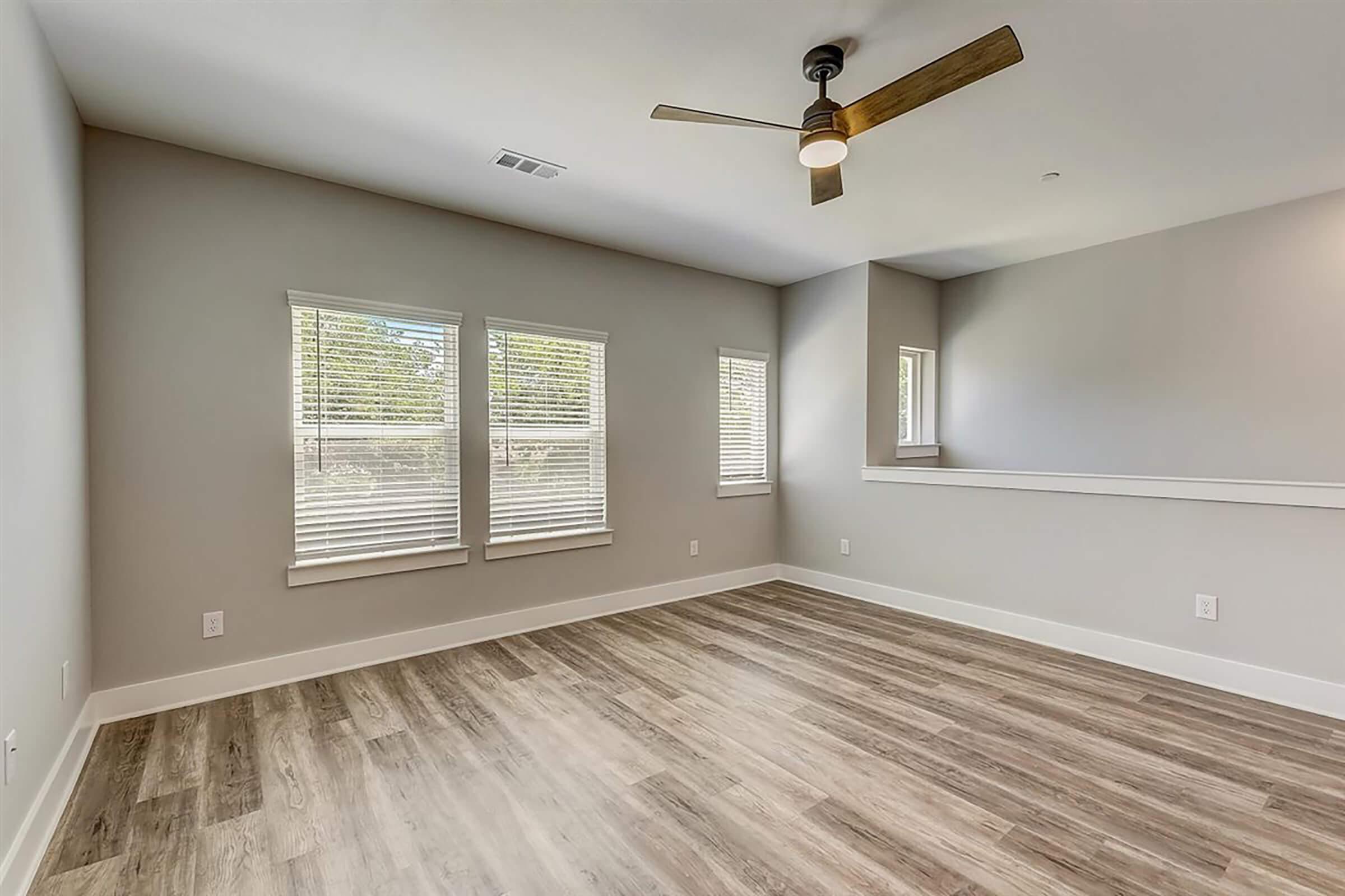 A spacious, well-lit living room featuring light wood flooring, large windows with white blinds, and a ceiling fan. The walls are painted in a neutral color, creating a modern and inviting atmosphere. There is a partial wall separating the space, possibly leading to another room or area.
