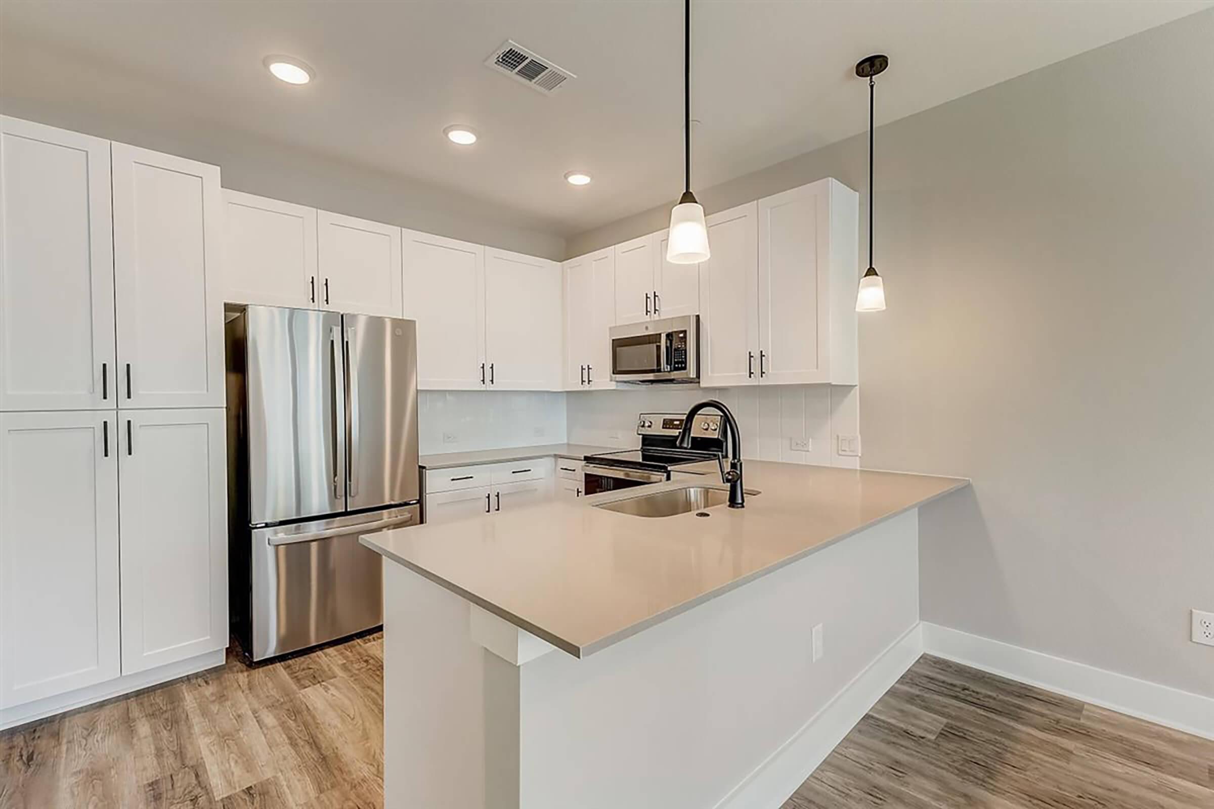 Modern kitchen featuring sleek white cabinetry, stainless steel appliances including a refrigerator and microwave, and a light-colored countertop. Pendant lighting hangs above the kitchen island, and the flooring has a warm wood-like finish, creating a bright and spacious atmosphere.