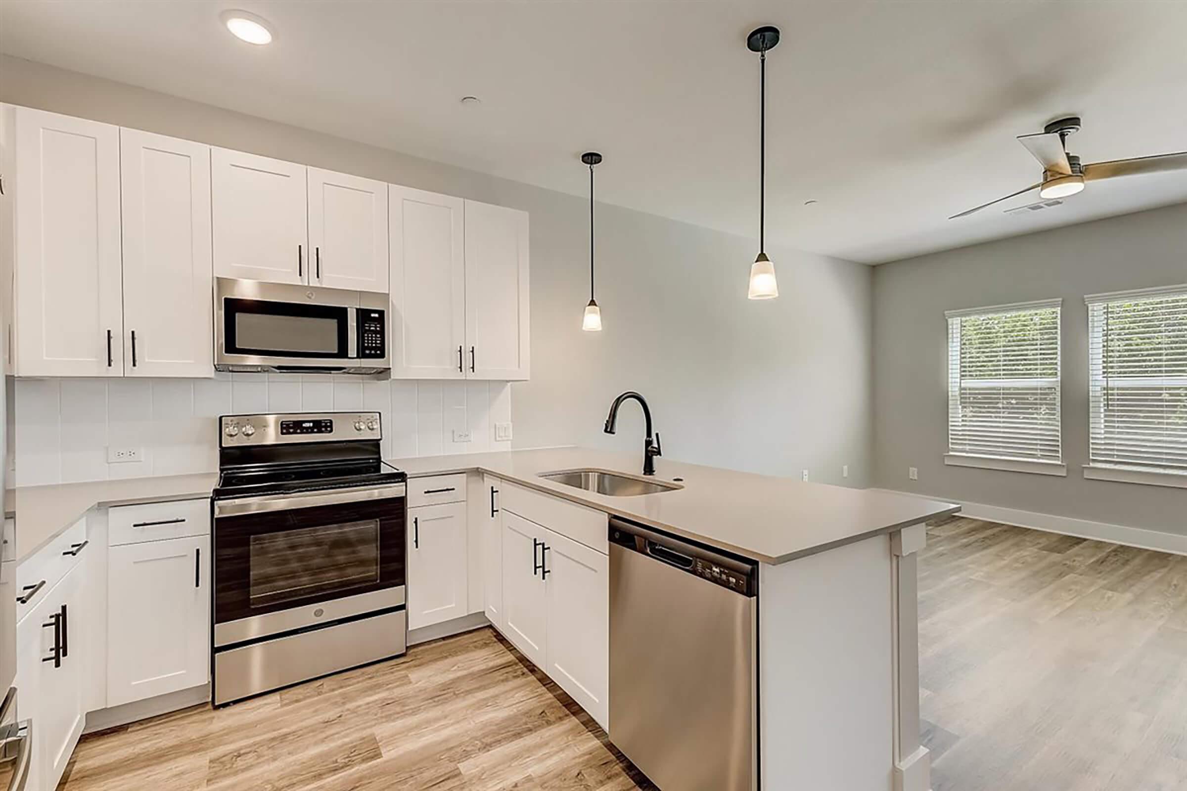 Modern kitchen featuring white cabinetry, stainless steel appliances including a microwave and oven, a sleek black faucet, and a dishwasher. The kitchen island has a countertop with bar seating. Pendant lighting hangs above, and there is a spacious living area visible with large windows for natural light.