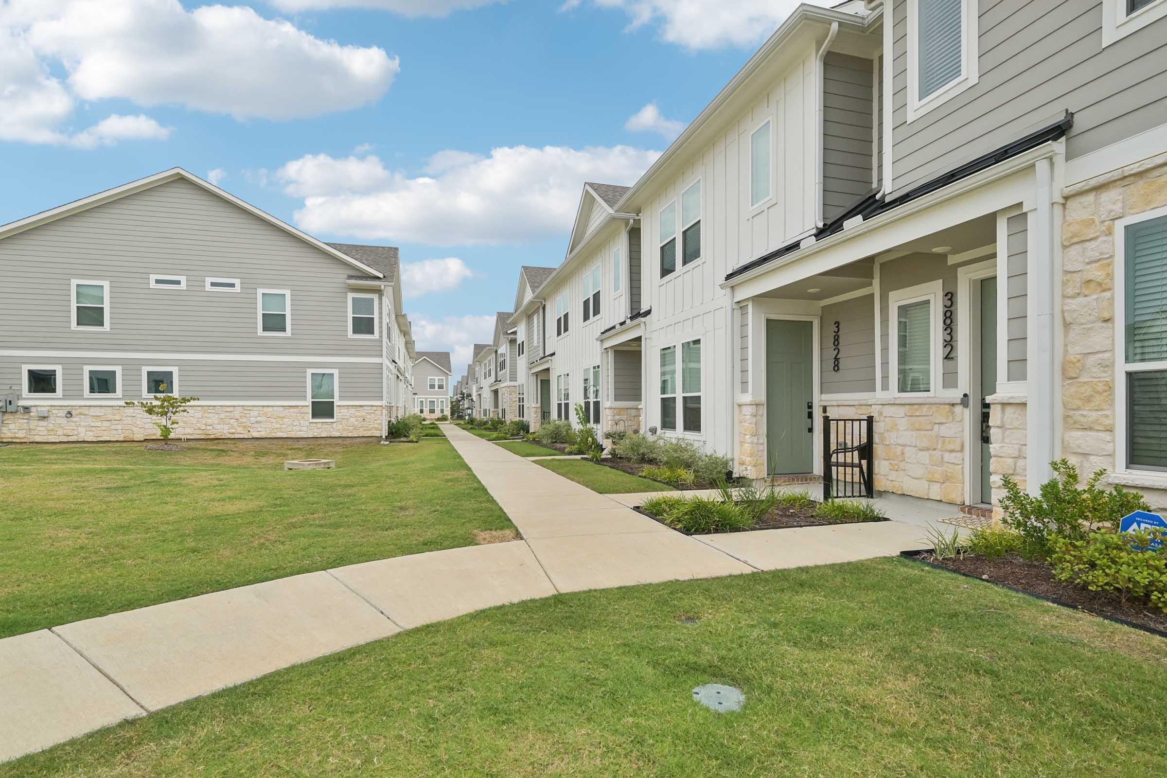 A row of modern townhouses featuring light-colored exteriors and stone accents, arranged along a paved walkway. The landscape includes well-maintained grass and small shrubs, with a partly cloudy blue sky overhead.