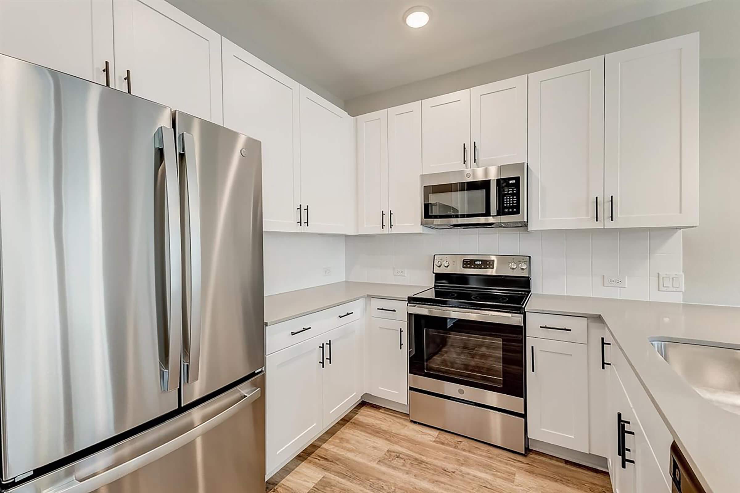 Modern kitchen featuring stainless steel appliances, including a refrigerator, oven, and microwave. The cabinetry is white with sleek handles, and the countertops are light-colored. A wooden floor adds warmth to the space, creating a bright and inviting atmosphere.