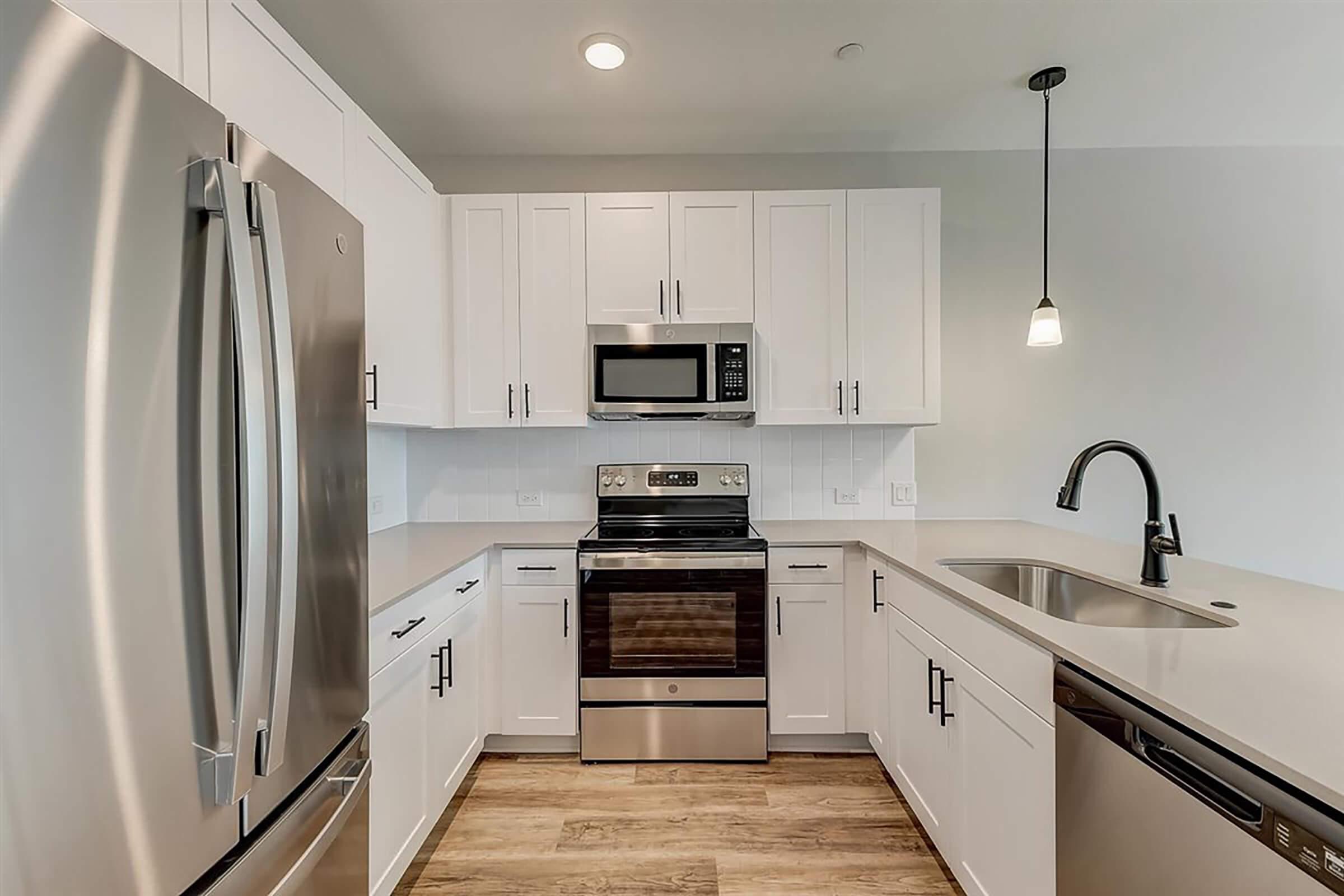 A modern kitchen featuring stainless steel appliances, including a refrigerator, stove, and microwave. The kitchen has white cabinets, a sleek countertop, and a single-basin sink with a pull-down faucet. A pendant light hangs above the counter, and the flooring is light wood.
