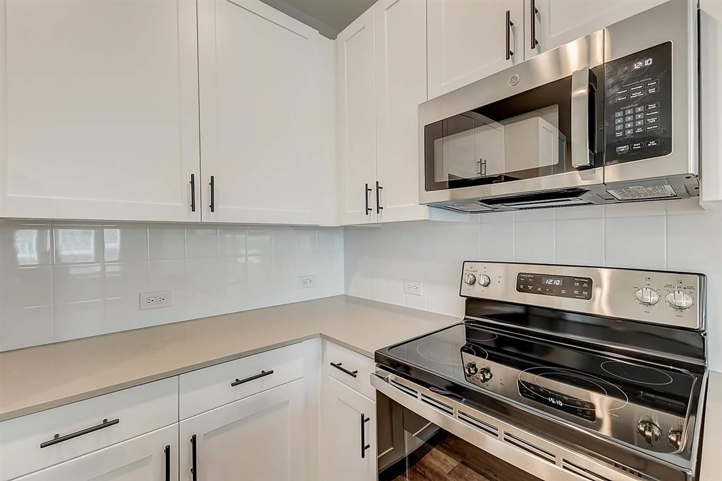 Modern kitchen featuring sleek white cabinetry with black handles, stainless steel microwave, and an oven. The countertop is a light neutral color, complementing the stylish design. Bright natural light illuminates the space, highlighting the minimalist aesthetic and clean lines.
