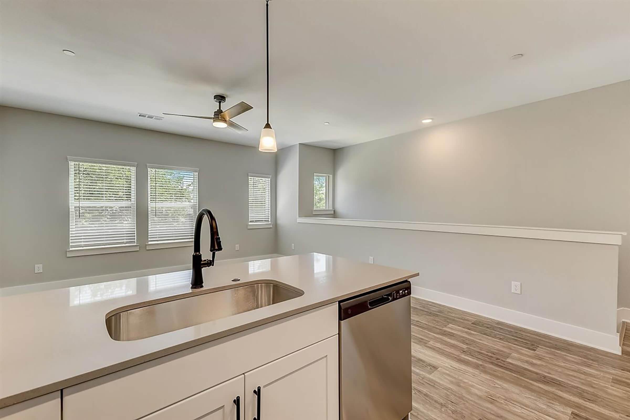 Modern kitchen with a sleek countertop, undermount sink, and stainless steel dishwasher. Bright and open living space in the background with large windows, allowing natural light to fill the area. Light-colored walls and wood-look flooring create a spacious and inviting atmosphere.