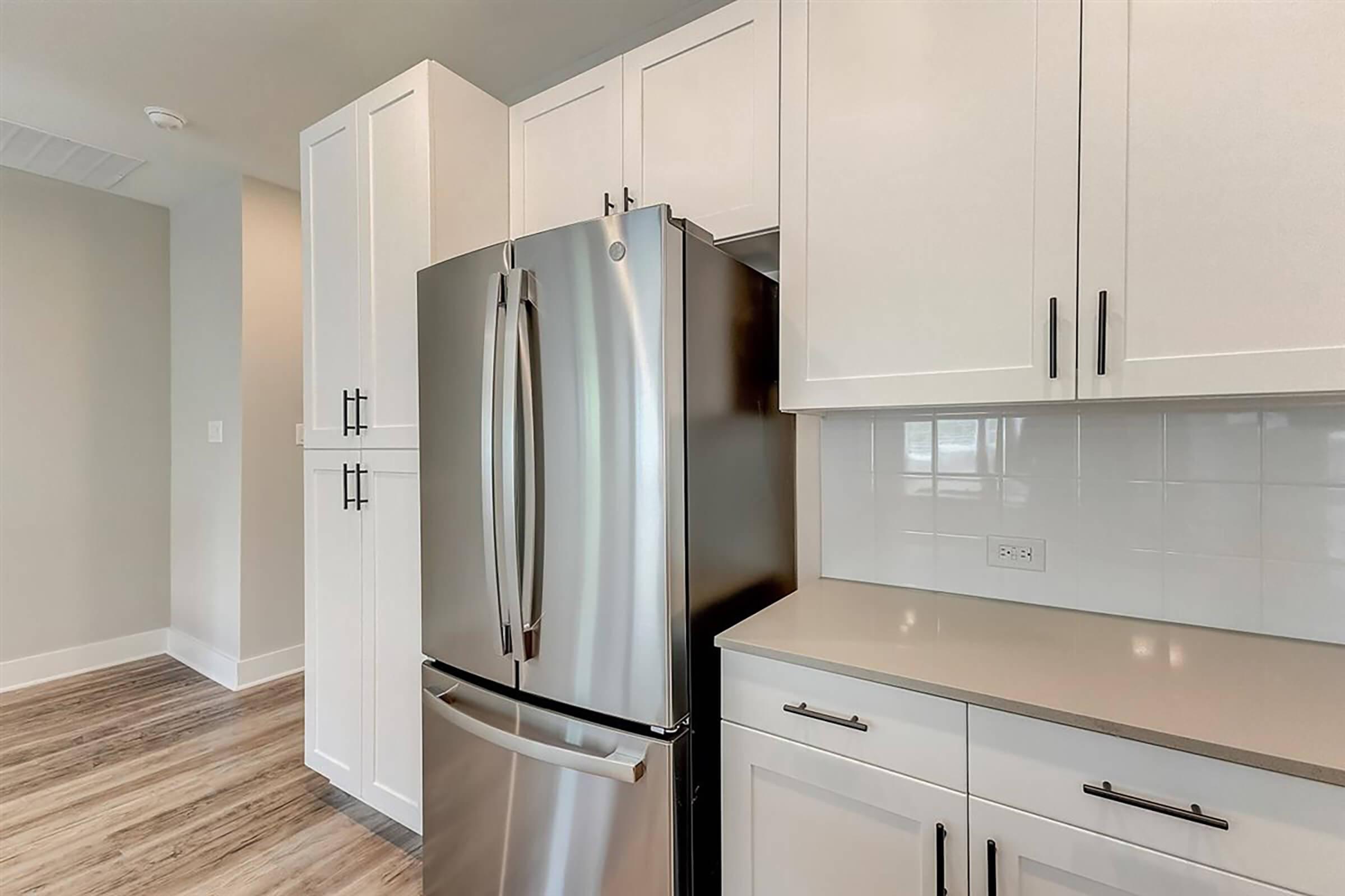 Modern kitchen featuring a stainless steel refrigerator next to white cabinetry with black handles. The countertop is light-colored, and the flooring is a wood-like laminate. The space is well-lit with natural light and has a neutral color scheme.