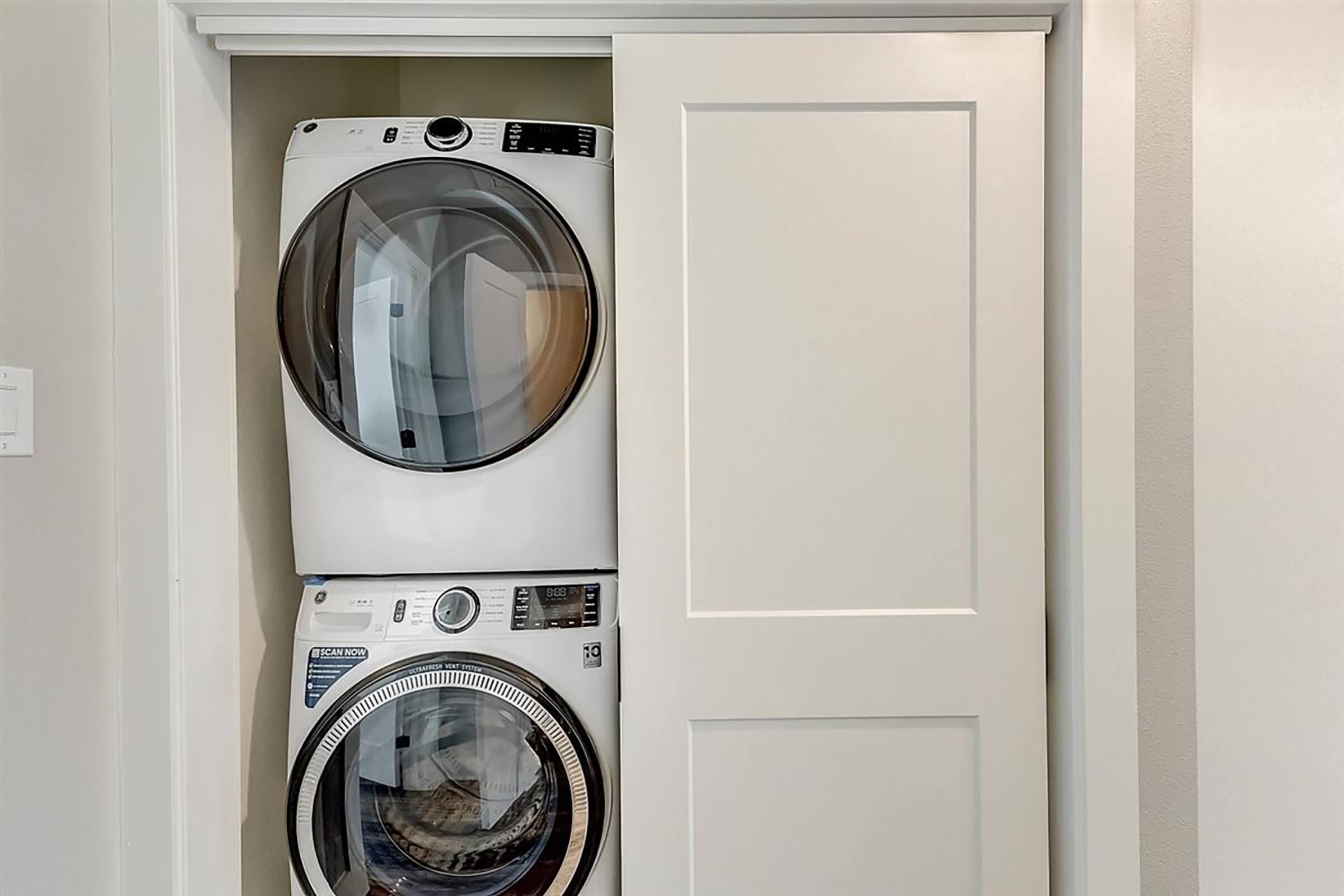 A modern laundry setup featuring a stacked washer and dryer within a closed white closet. The appliances have a sleek design with digital controls, and the closet door is partially opened, revealing the machines inside.