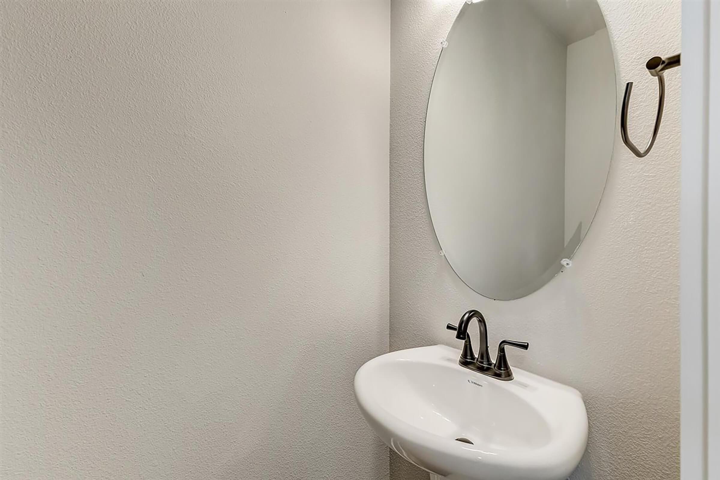 A small bathroom featuring a round mirror above a white sink with two faucets. The walls are painted a light color, and there is a simple wall hook beside the mirror. The space is well-lit and uncluttered, creating a clean and minimalist look.