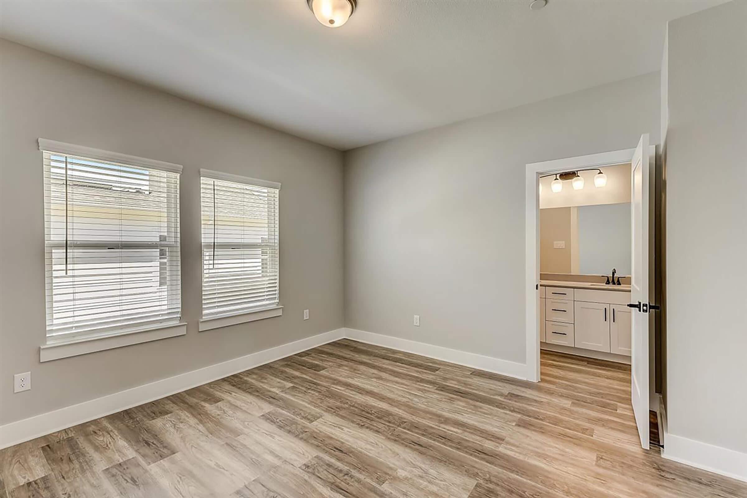 A spacious, well-lit room featuring two windows with blinds, light hardwood flooring, and neutral-colored walls. To the right, there's a doorway leading to a bathroom with visible lighting fixtures and modern fittings. The overall atmosphere is clean and inviting, ideal for various uses.