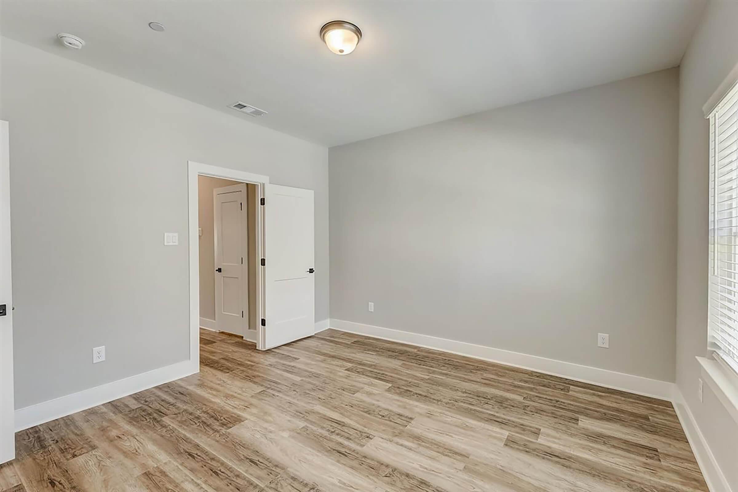 A spacious, empty room featuring light gray walls, wooden flooring, and bright natural light from a window. The room includes a closed door on the left and an open door leading to a hallway on the right, with a ceiling light fixture in the center.