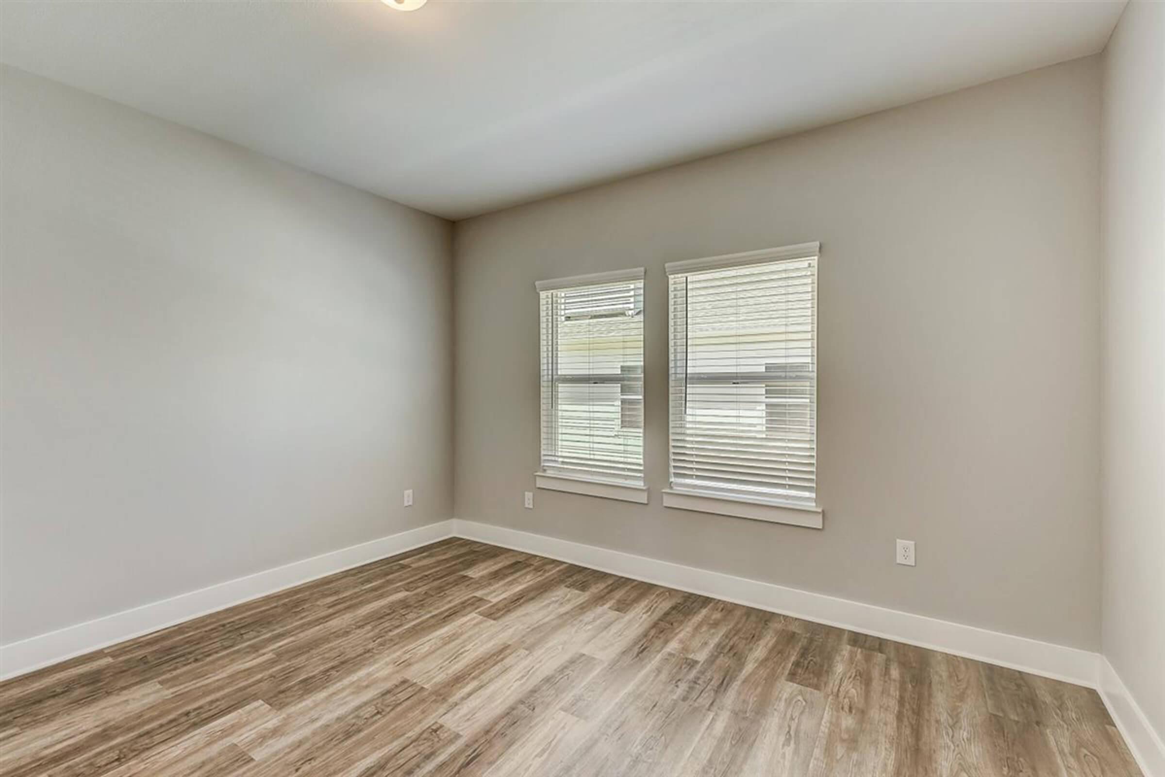 A bright, empty room with light-colored walls and large windows featuring blinds. The floor is made of light wood laminate, and there is a soft ceiling light. The space appears spacious and ready for furnishing.