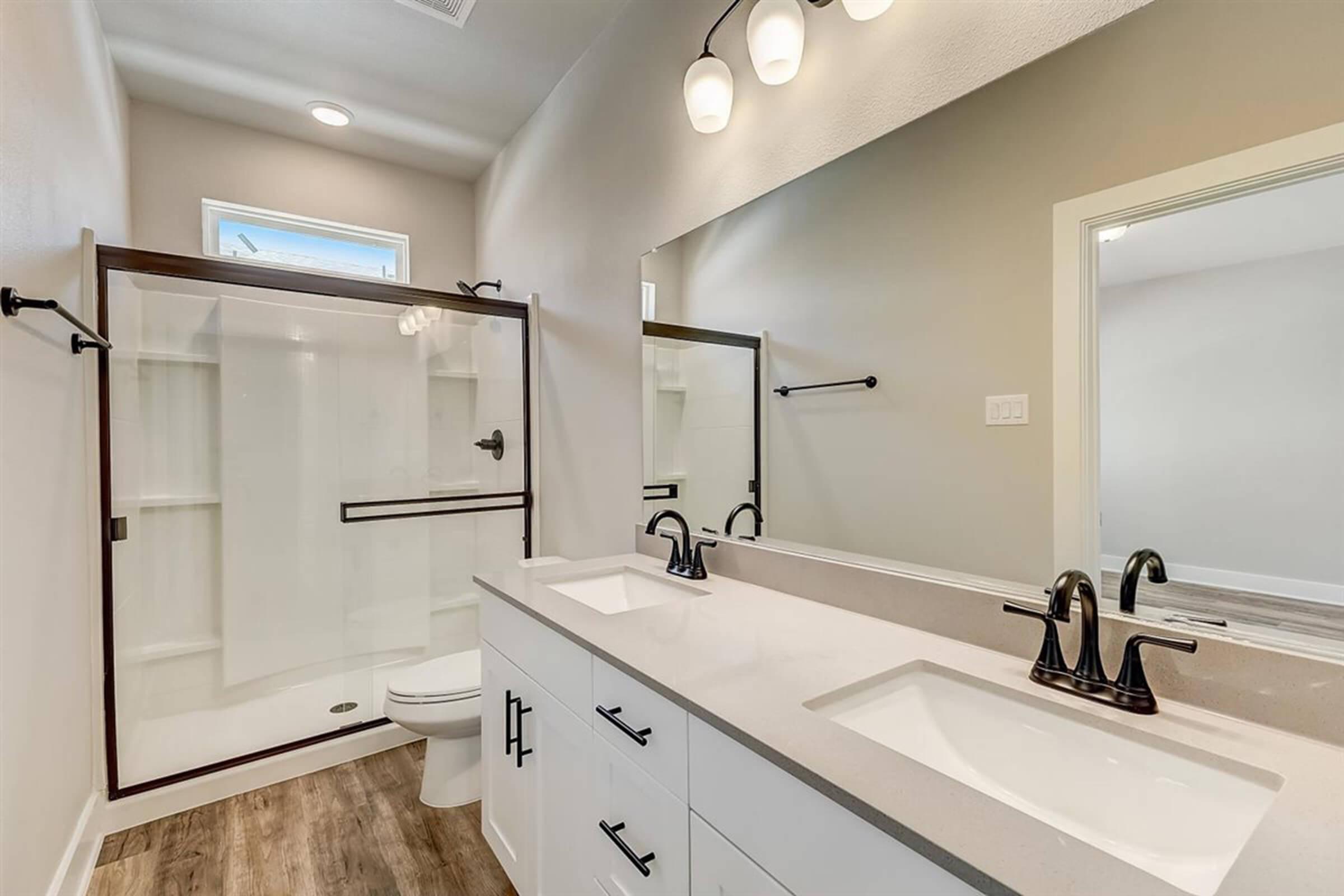 A modern bathroom featuring a double sink vanity with black faucets, a large mirror, and a walk-in shower with glass doors. The walls are painted in a light gray color, and the flooring is a wood-like laminate. Natural light enters through a window above the shower.