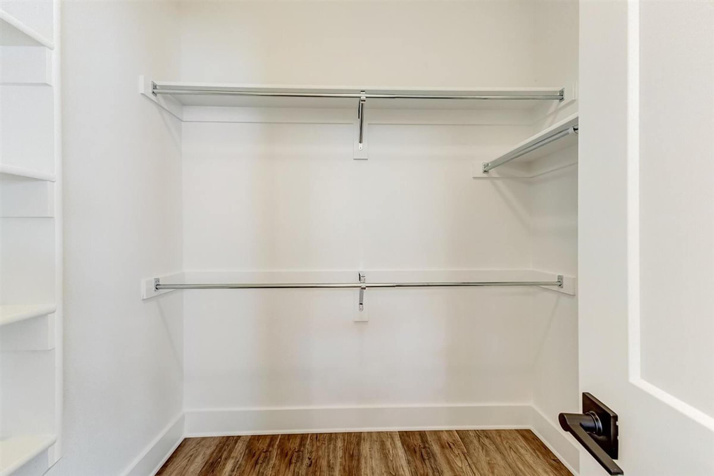 An empty closet with white walls and a light wooden floor. It features two horizontal shelves mounted on the walls, and a door handle visible on the right side. The space is uncluttered, creating a minimalist look.