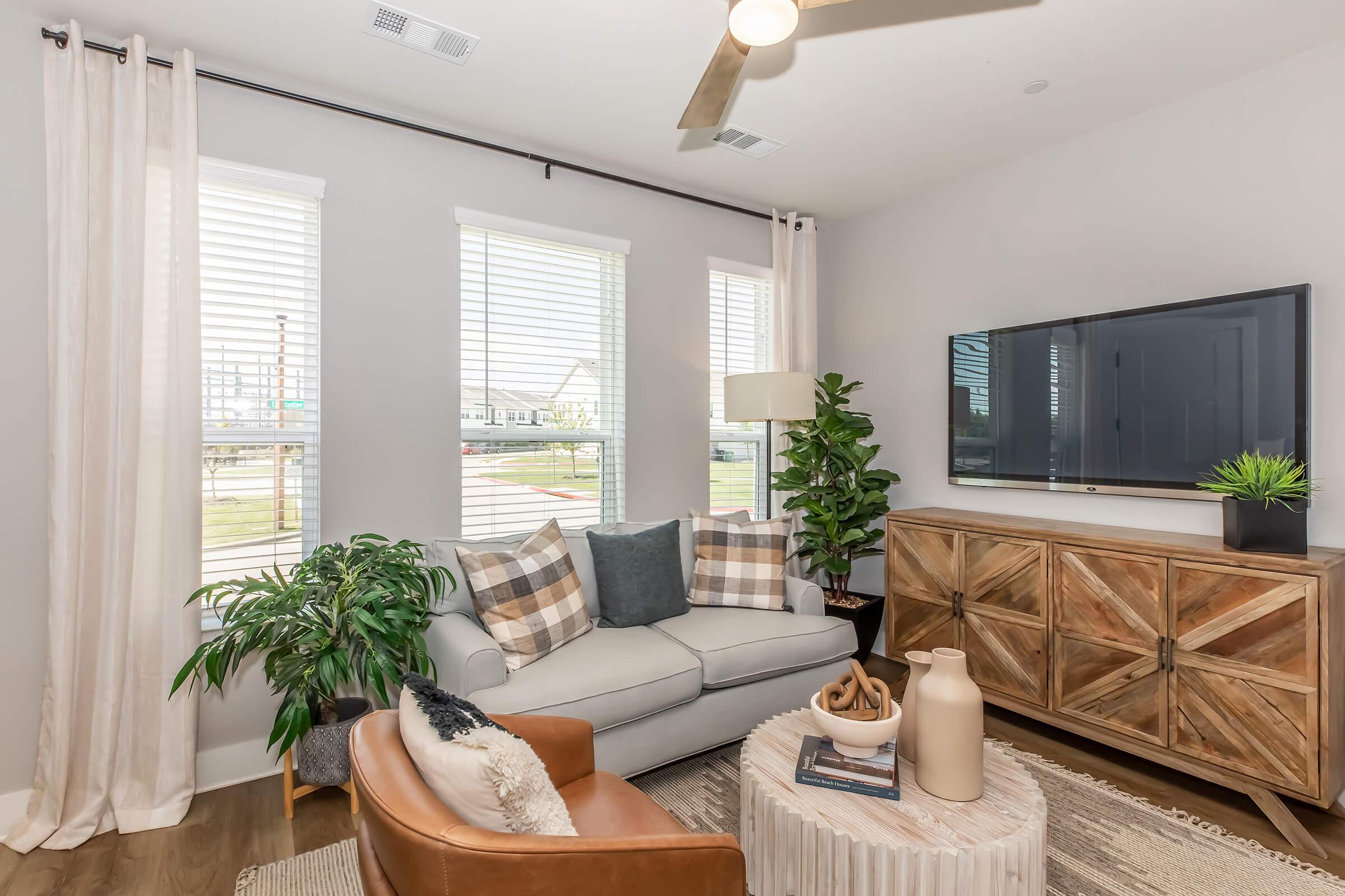 A cozy living room featuring a light gray sofa with decorative pillows, a tan leather chair, and a wooden side table. Natural light streams in through three large windows with sheer curtains, and a flat-screen TV is mounted on the wall. A stylish cabinet and indoor plants add warmth to the space.