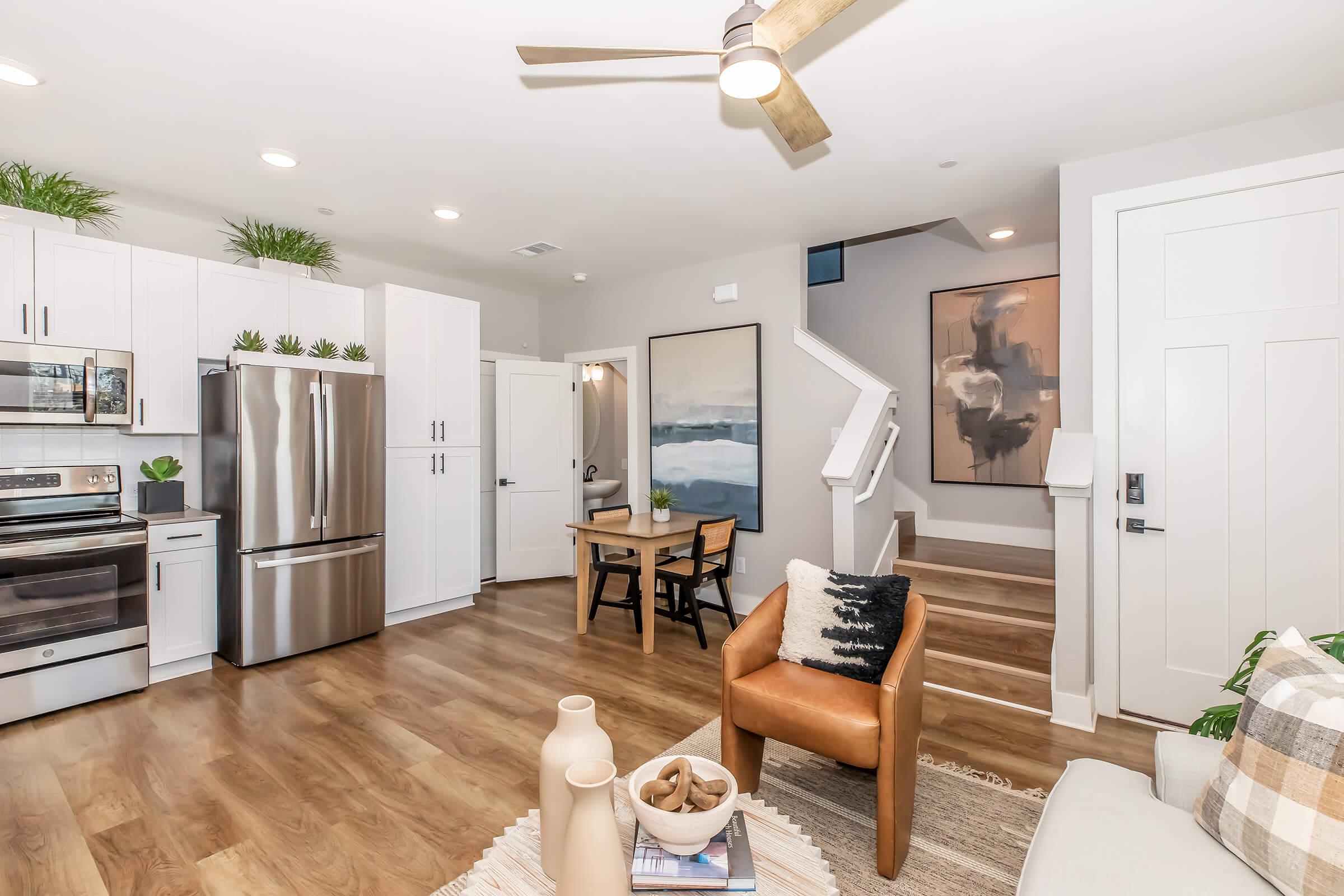 Modern living room and kitchen area featuring stainless steel appliances, white cabinetry, and a wooden table with chairs. There is a cozy seating area with a brown armchair and decorative items on a table. A staircase leads to an upper level, and the space is enhanced with plants and contemporary artwork.