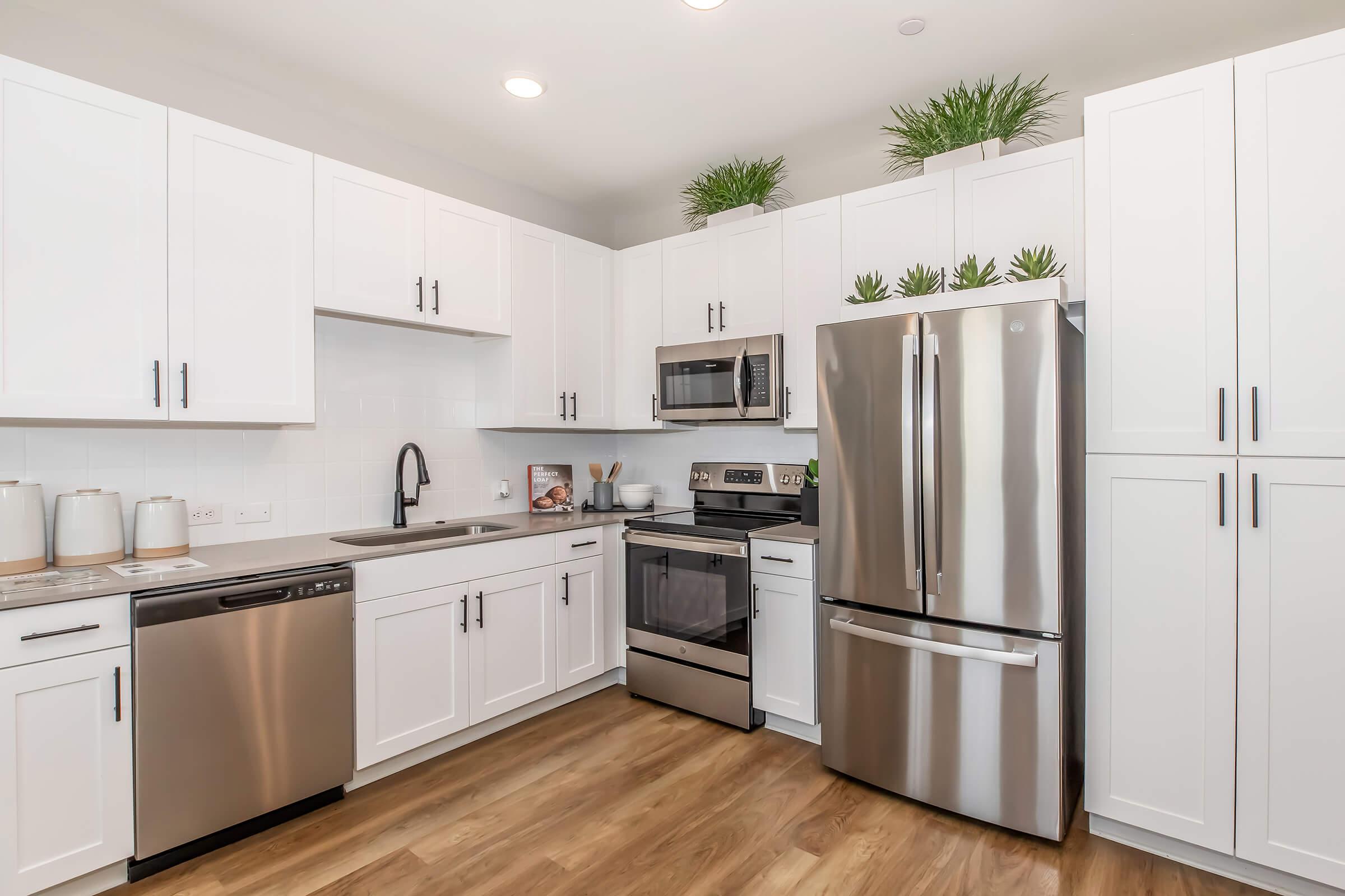 Modern kitchen featuring white cabinetry, stainless steel appliances including a refrigerator, microwave, and oven. The countertop is light-colored, and a sink is located next to the dishwasher. Bright lighting illuminates the space, complemented by small potted plants on top of the cabinets. Hardwood flooring completes the design.