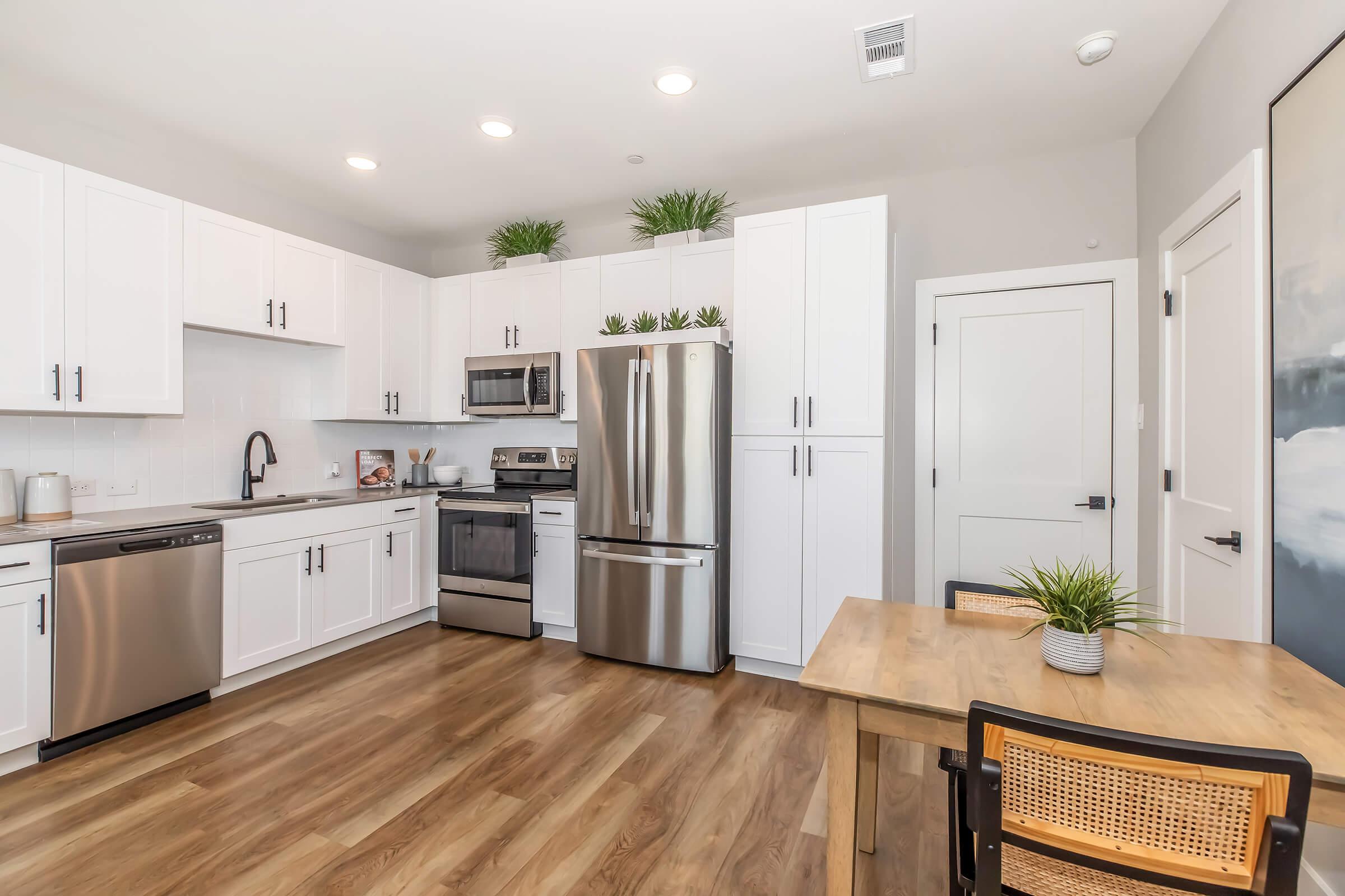 Modern kitchen featuring white cabinetry and stainless steel appliances, including a refrigerator and microwave. The space includes a wooden dining table with a plant centerpiece, hardwood flooring, and bright overhead lighting. Simple decor with green plants on the cabinets adds a fresh touch.