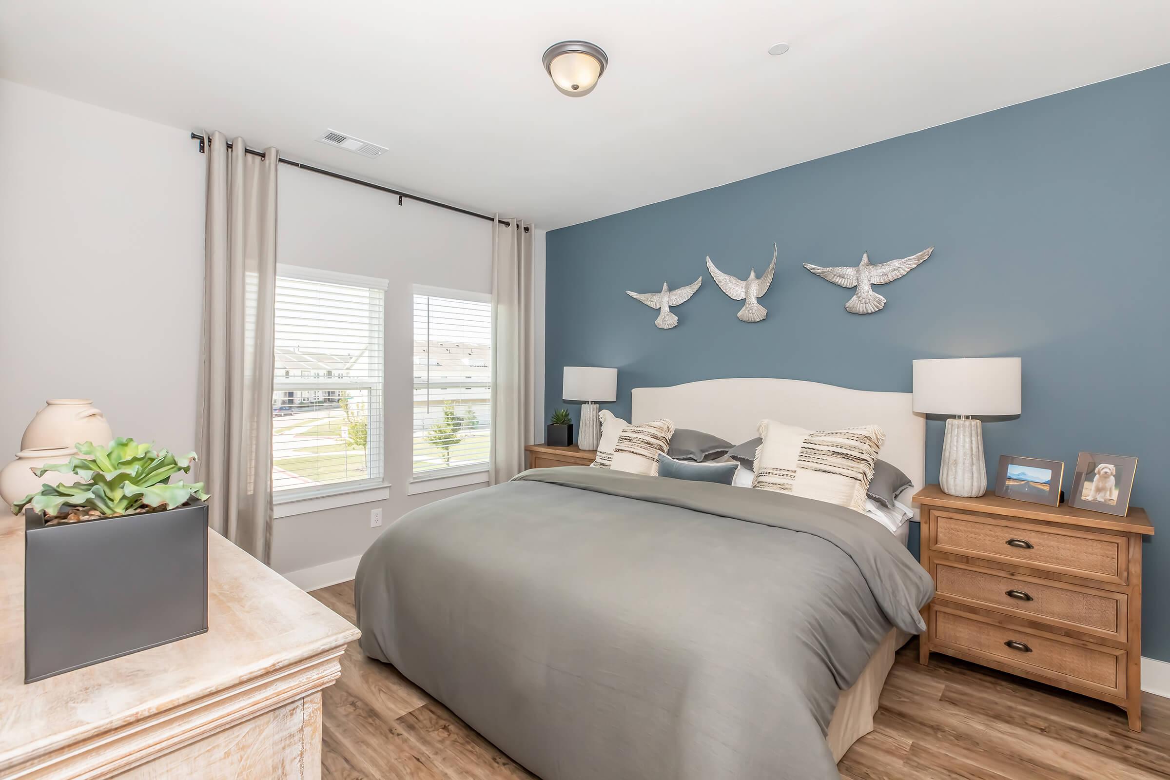 Cozy bedroom featuring a king-sized bed with gray bedding, two bedside tables with lamps, and a decorative wall with three bird sculptures. Light-colored curtains frame a window, letting in natural light, while a plant adds a touch of greenery to the room's modern decor.