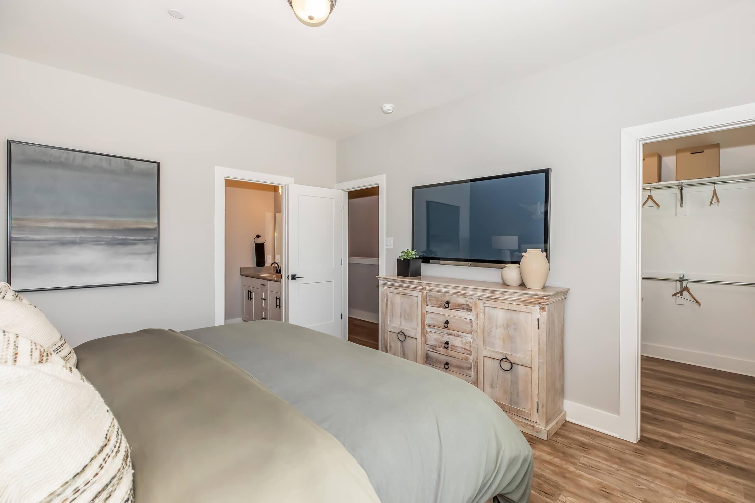 A neatly arranged bedroom featuring a large bed with a gray comforter, a wooden dresser with decorative items, a mounted flat-screen TV, and light-colored walls. A bathroom entrance and a closet are visible in the background, creating a modern and inviting atmosphere.