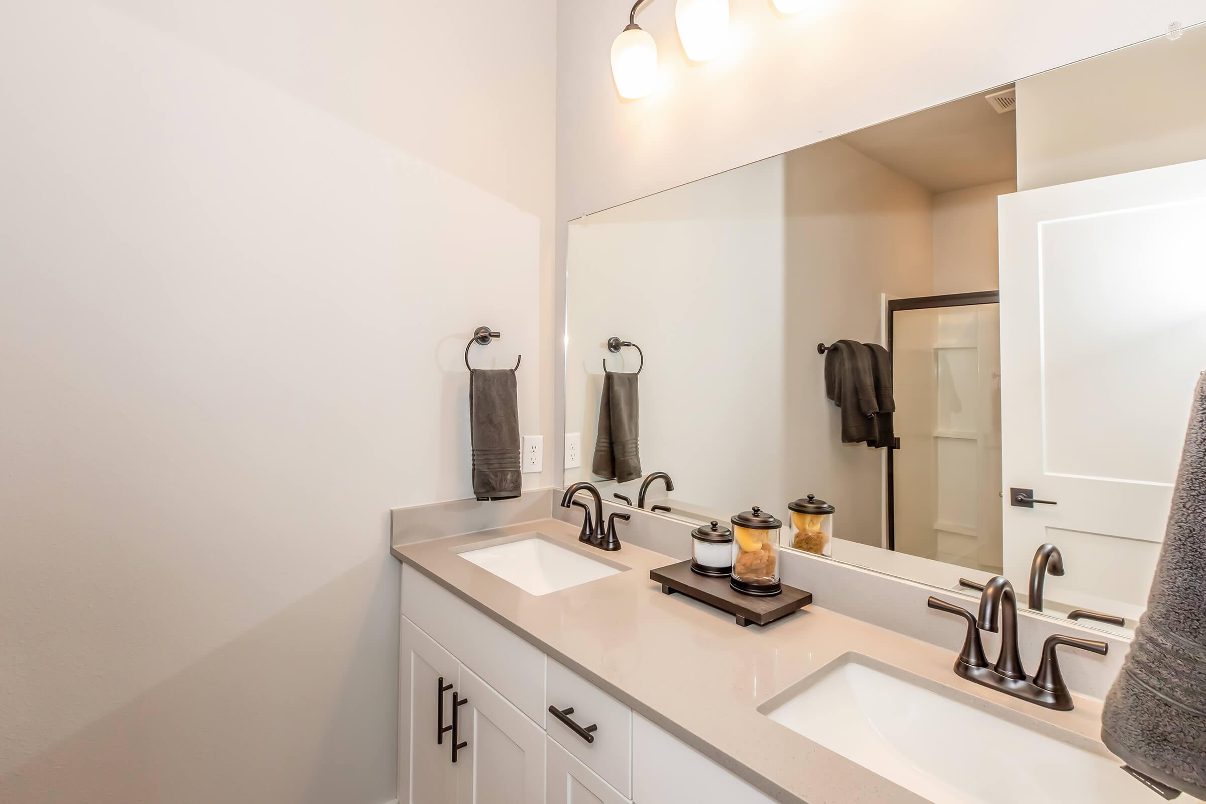 Modern bathroom featuring a double vanity with two sinks, stylish faucet fixtures, and a large mirror above. The countertop is light-colored with decorative items, including a small tray with jars. Two hand towels are hanging on wall-mounted racks, and a shower is visible behind a frosted glass door.