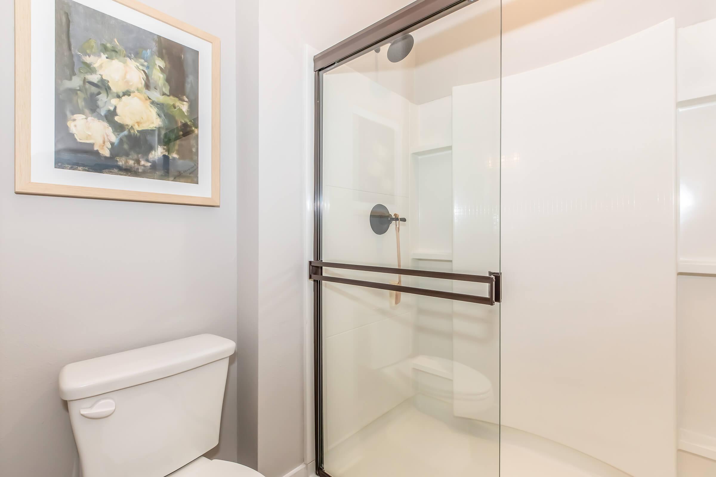 A modern bathroom featuring a walk-in shower with a glass door, a white toilet, and a framed painting of flowers on the wall. The walls are painted in a light color, creating a clean and bright atmosphere.