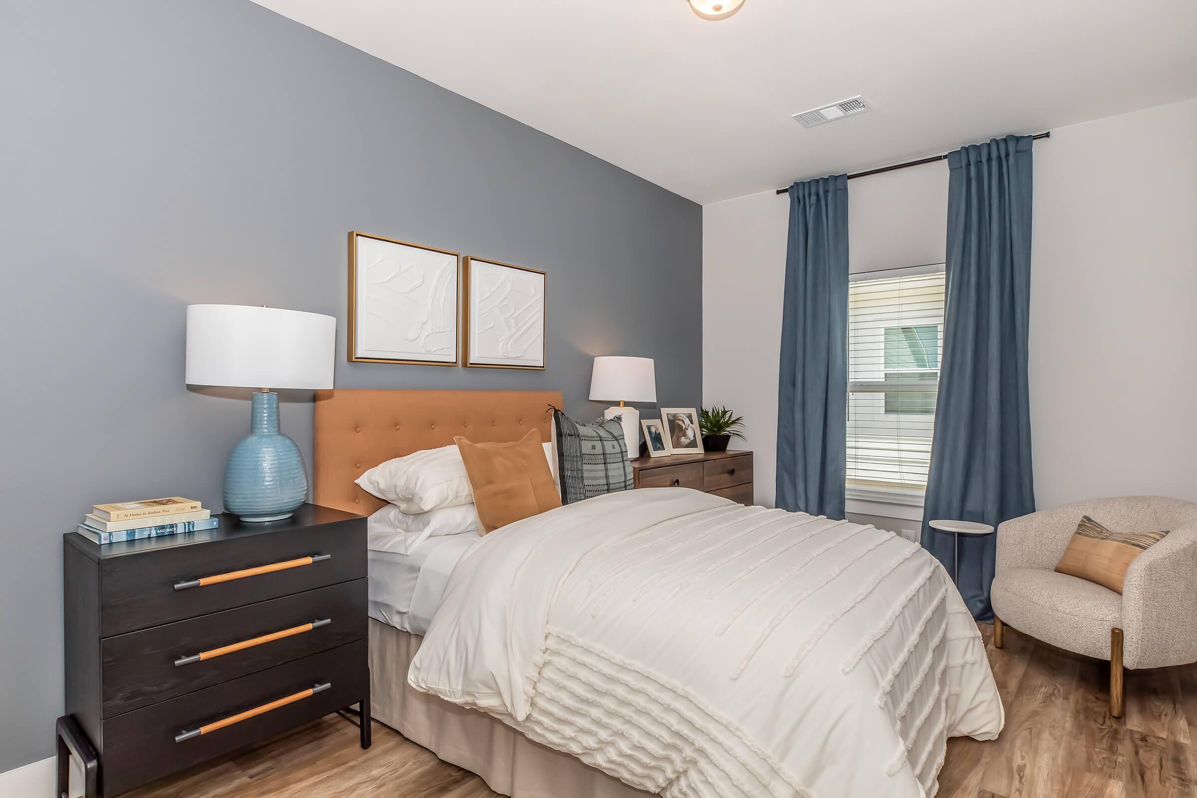 A cozy bedroom featuring a beige bed with white bedding and a tan headboard, flanked by two stylish lamps. A dark wood nightstand holds decorative items, and a light gray armchair is positioned nearby. The walls are painted blue, and there are two framed artworks above the bed and blue curtains on the window.