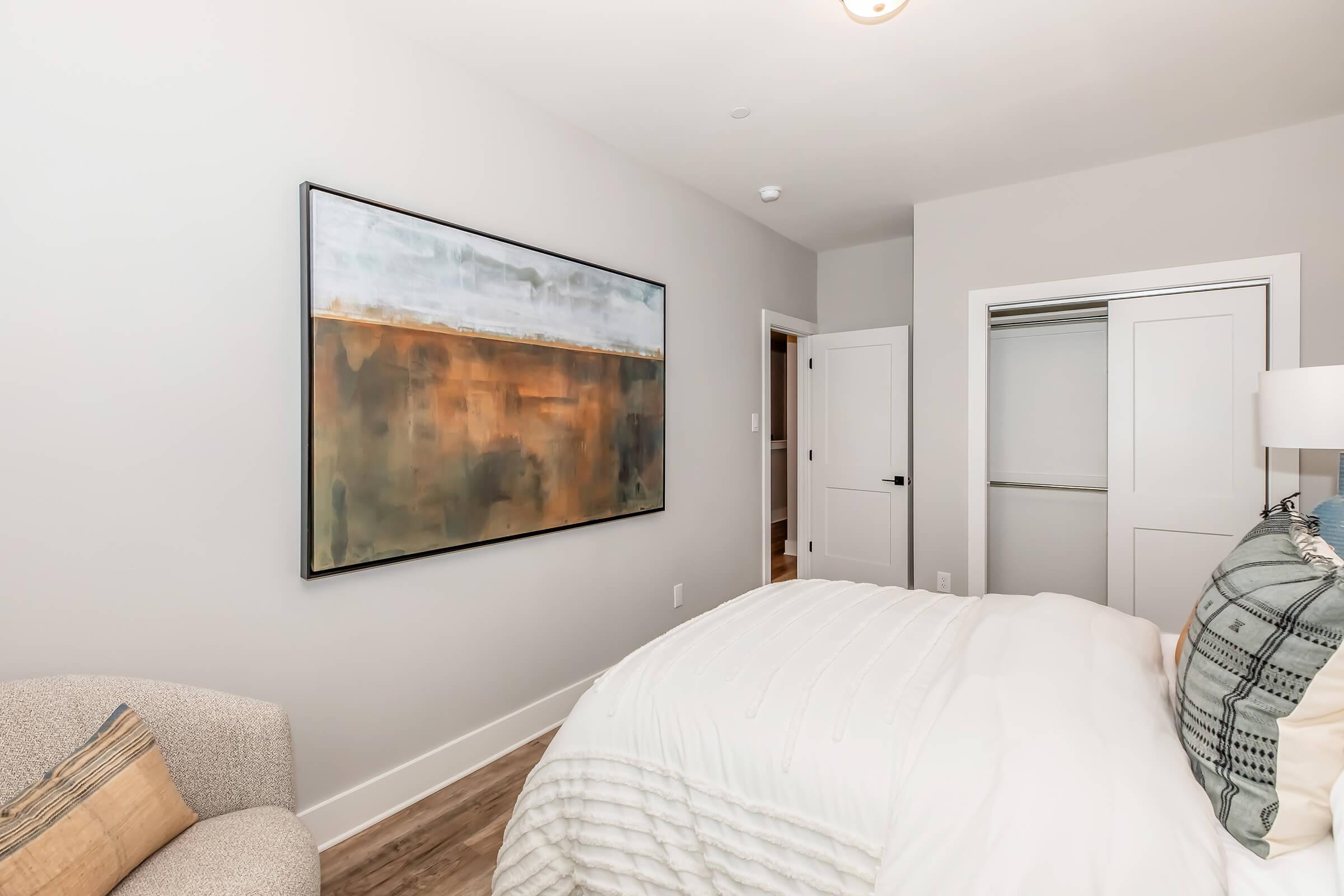 A modern bedroom featuring a comfortable white bed with decorative pillows, a light gray armchair, and a large abstract painting on the wall that incorporates earthy tones. A closet is visible in the corner, with a soft, neutral-colored wall and wooden flooring completing the design.