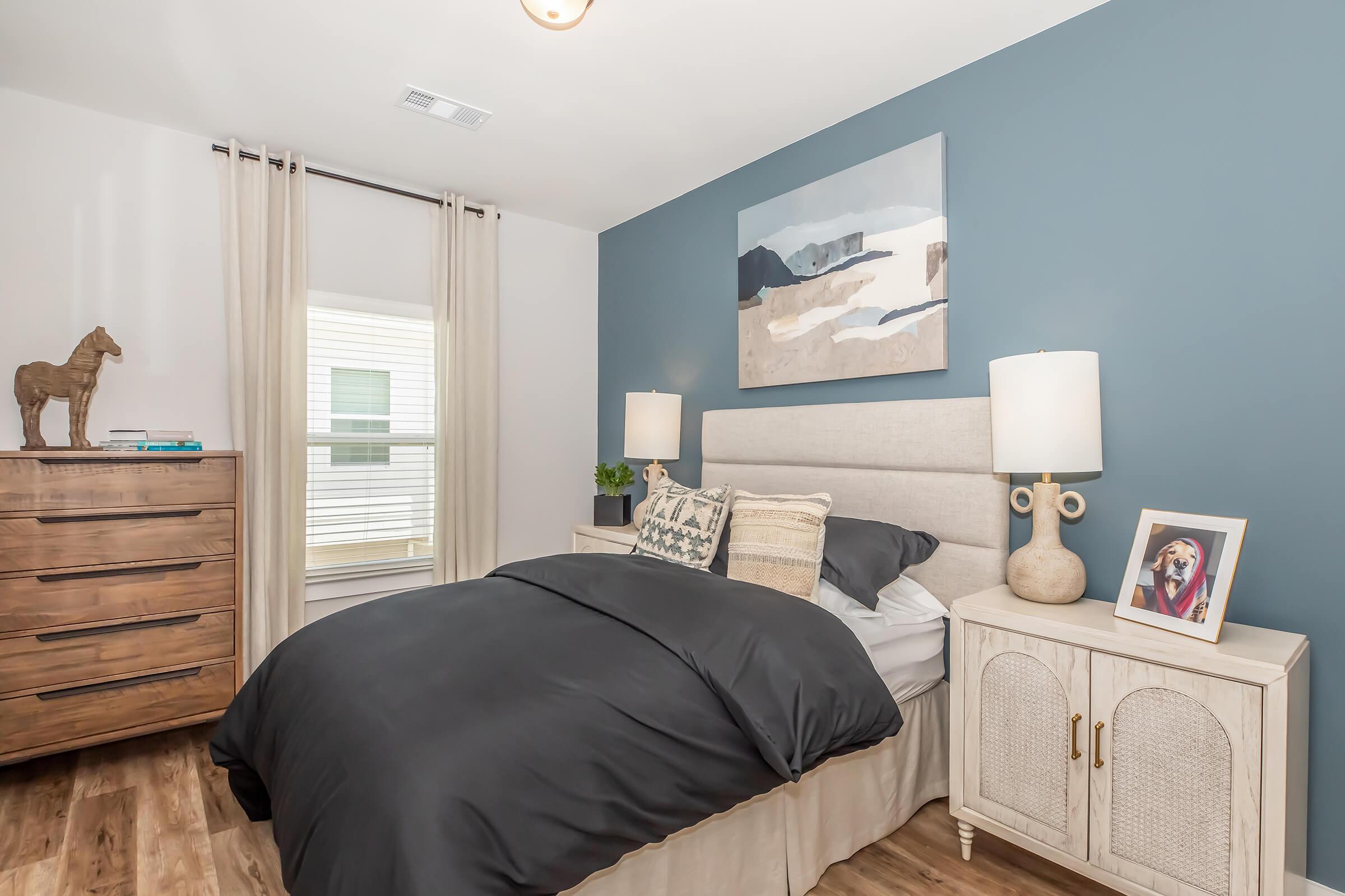 A cozy bedroom featuring a gray and white color scheme. The bed is dressed in a dark comforter, complemented by light gray pillows. A wooden dresser with a horse figurine sits nearby, and a framed photo is displayed on a nightstand. Soft curtains frame a window, allowing natural light to brighten the space.