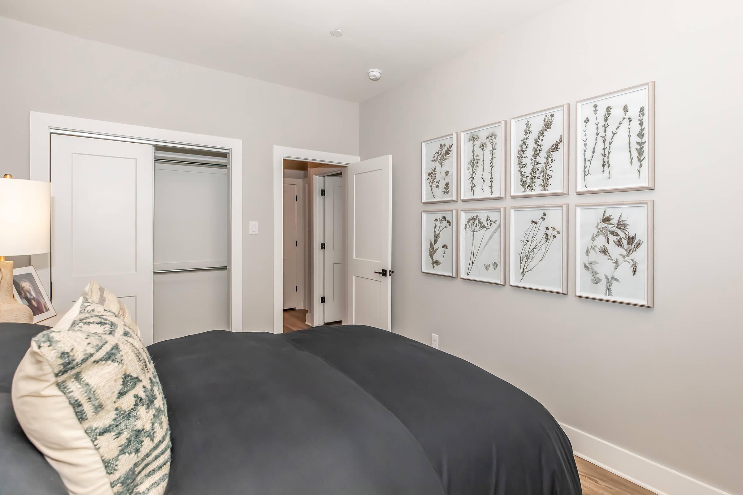A modern bedroom featuring a dark bedspread and a decorative pillow. On the wall, there are eight framed botanical prints. A closet door is partially visible, along with a doorway leading to another room, all set against neutral-toned walls and wooden flooring.