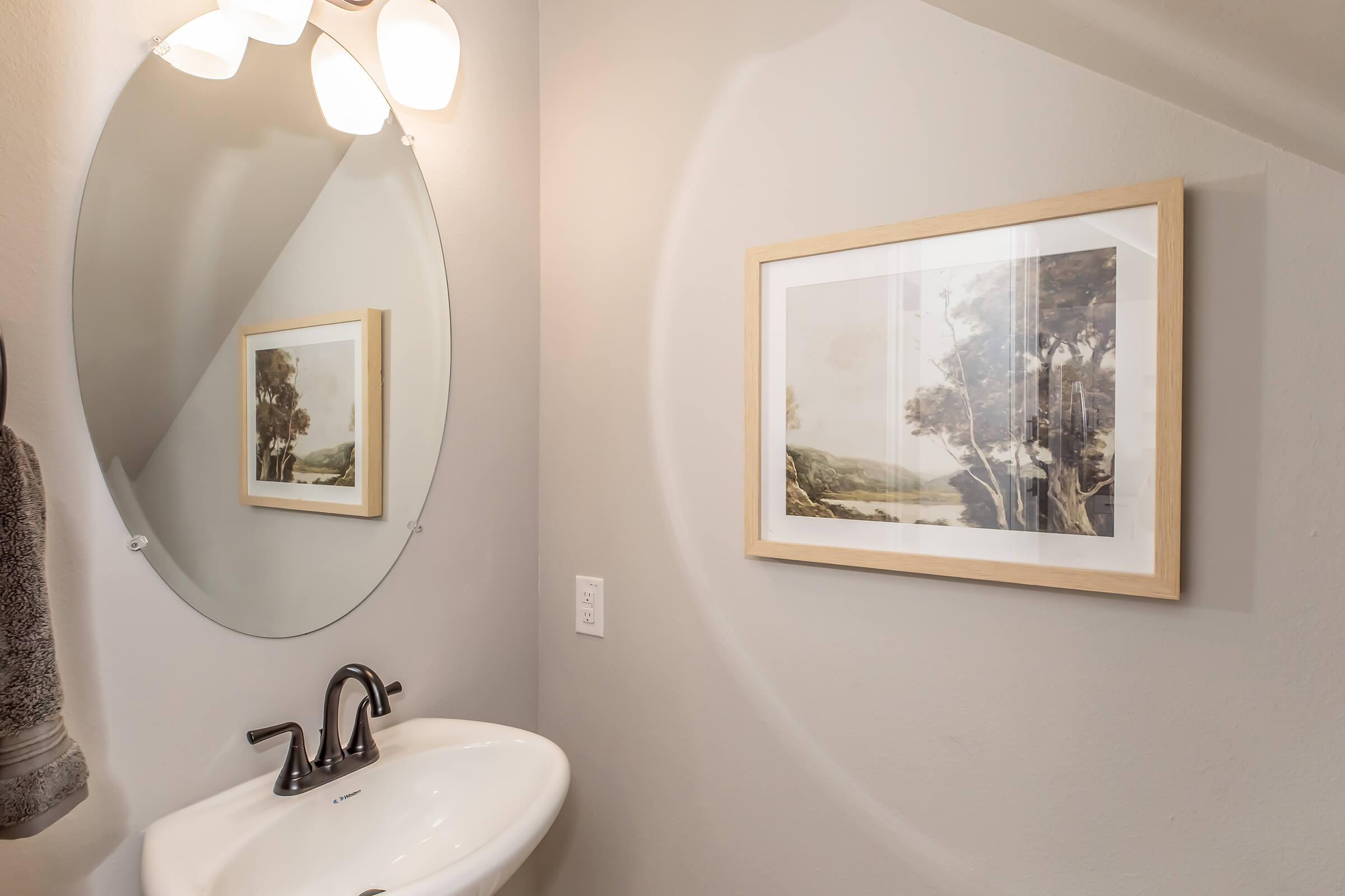 A modern bathroom featuring a circular mirror above a white sink. The walls are painted in a soft gray color. A framed artwork hangs on the wall, depicting a landscape scene with trees and mountains. A towel is neatly hung nearby, adding to the serene atmosphere.