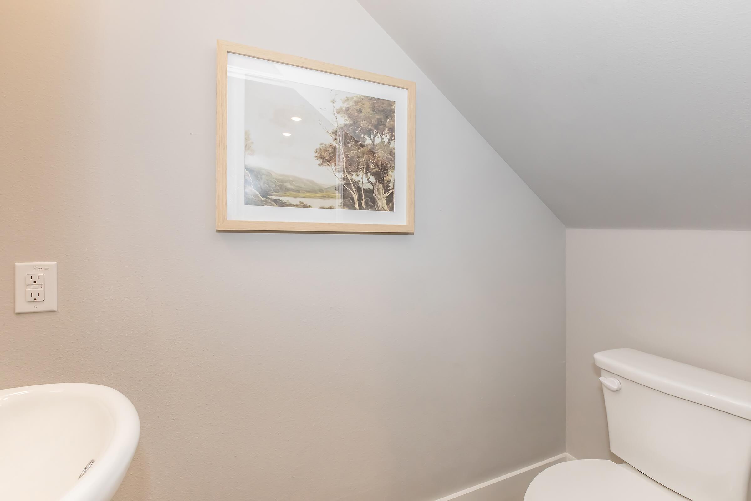 A bathroom scene featuring a framed landscape painting on the wall, with a toilet visible nearby. The walls are painted in a light gray color, and the ceiling slopes slightly, creating a cozy atmosphere. A light switch is located beside the painting.