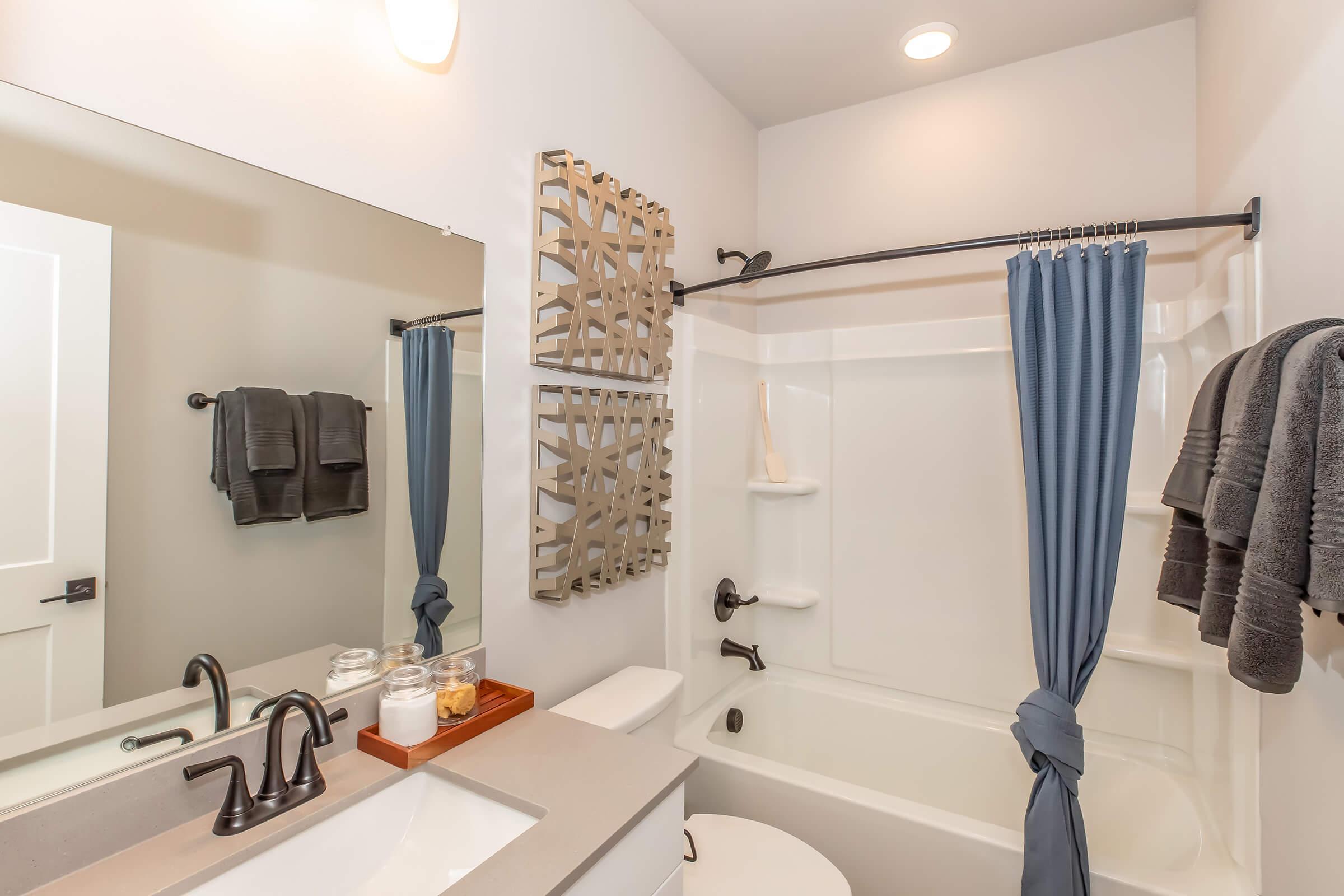 A modern bathroom featuring a white bathtub with a blue curtain, a sleek gray countertop with a sink, and a mirror. Wall decor includes decorative panels, and there are gray towels hanging on a rack. The space is bright with neutral tones and contemporary fixtures.