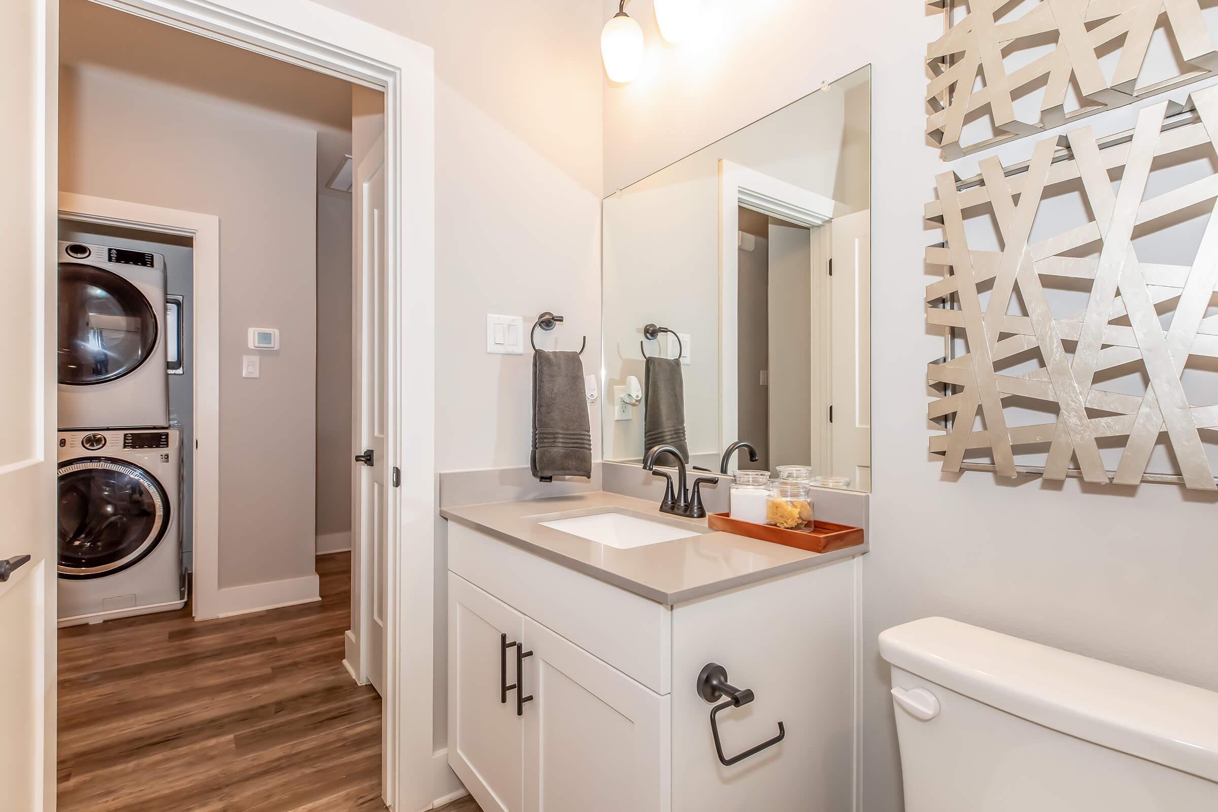 Modern bathroom featuring a sleek vanity with a sink and a large mirror, complemented by decorative wall art. A stackable washer and dryer is visible through an open door, and the space has warm wood-like flooring and neutral wall colors for a contemporary feel.
