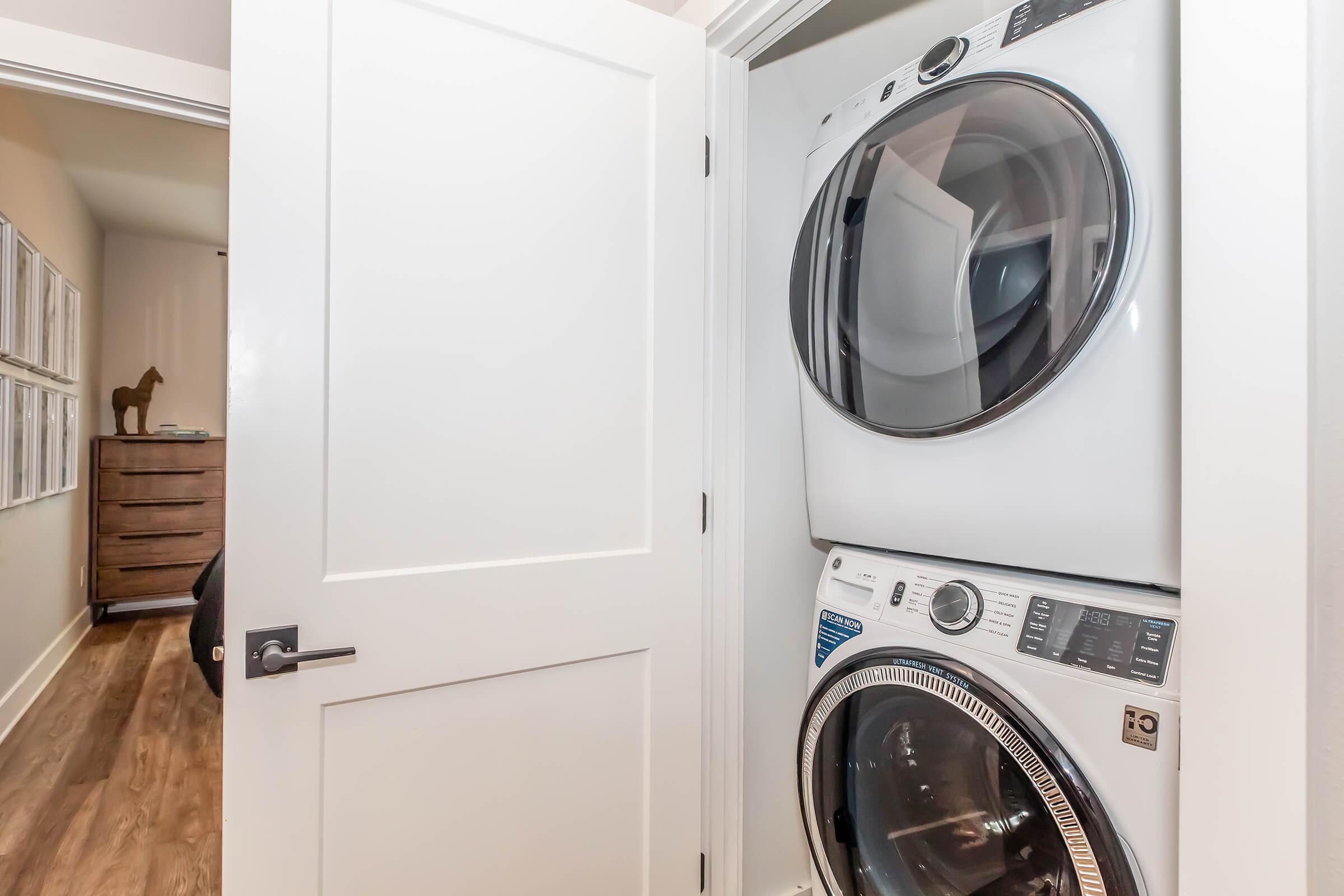 A compact laundry area featuring a stacked washer and dryer within a closed door setting. The background includes a glimpse of a dresser and a subtle decorative element, contributing to a modern, organized home environment. The flooring is hardwood, enhancing the aesthetic.