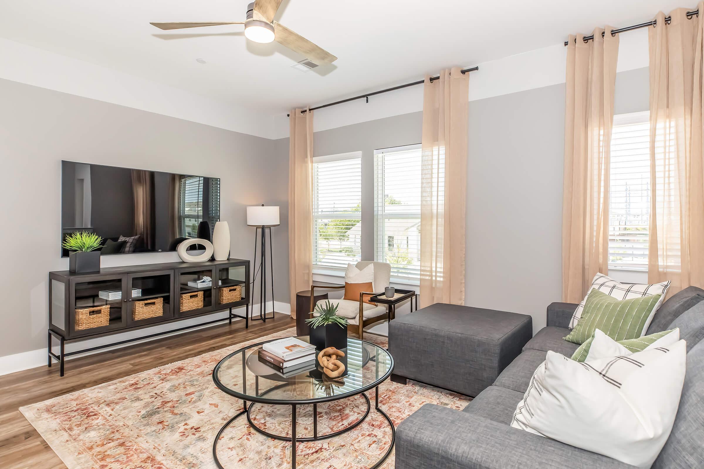 A modern living room featuring a gray sectional sofa, a round glass coffee table, and a black television cabinet. Decorative items include potted plants and a few books. Natural light filters through sheer curtains, enhancing the inviting atmosphere. Wood-like flooring adds warmth to the space.
