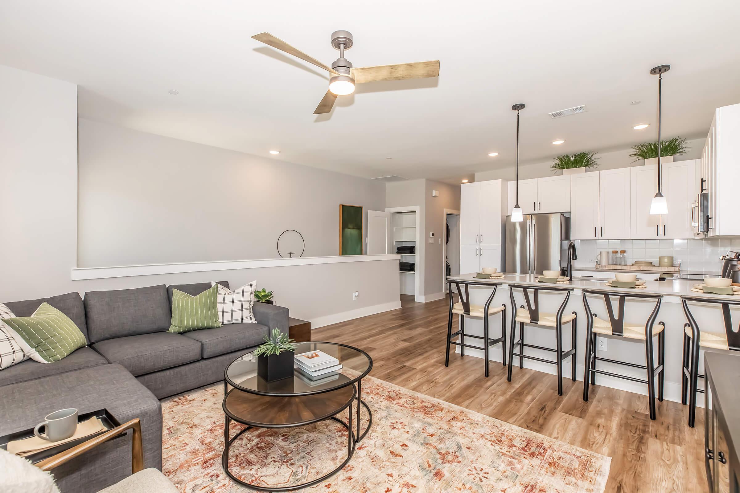Modern open-concept living space featuring a gray sectional sofa, a round coffee table, and hardwood flooring. The kitchen includes white cabinetry, stainless steel appliances, and bar seating with stylish pendant lighting above. A decorative area rug adds warmth to the room.