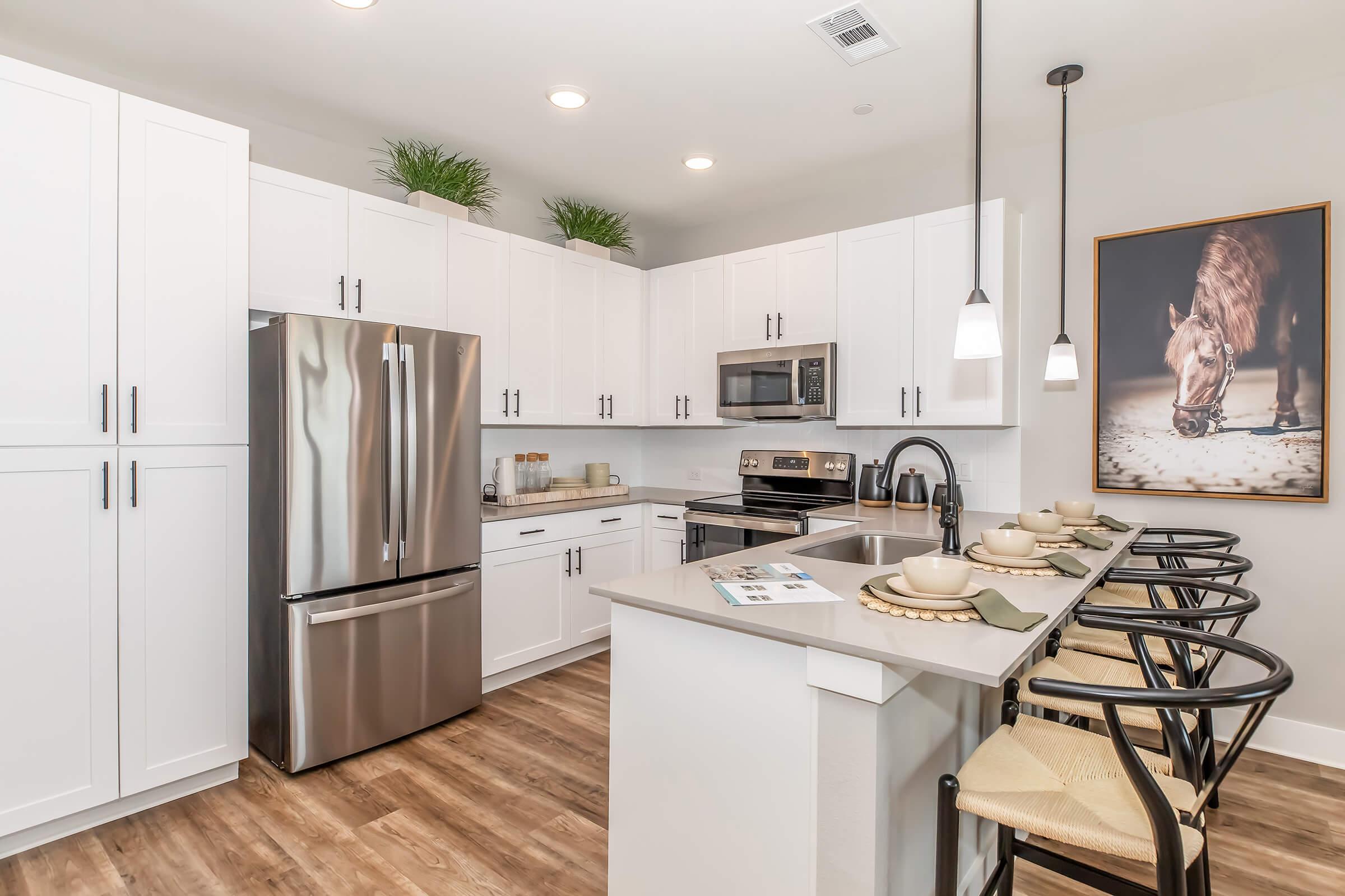 A modern kitchen featuring white cabinetry, stainless steel appliances, and a large island with barstools. The space is well-lit and includes decorative plants, a horse-themed artwork on the wall, and an organized dining area with plates and utensils. The flooring is wood-like.