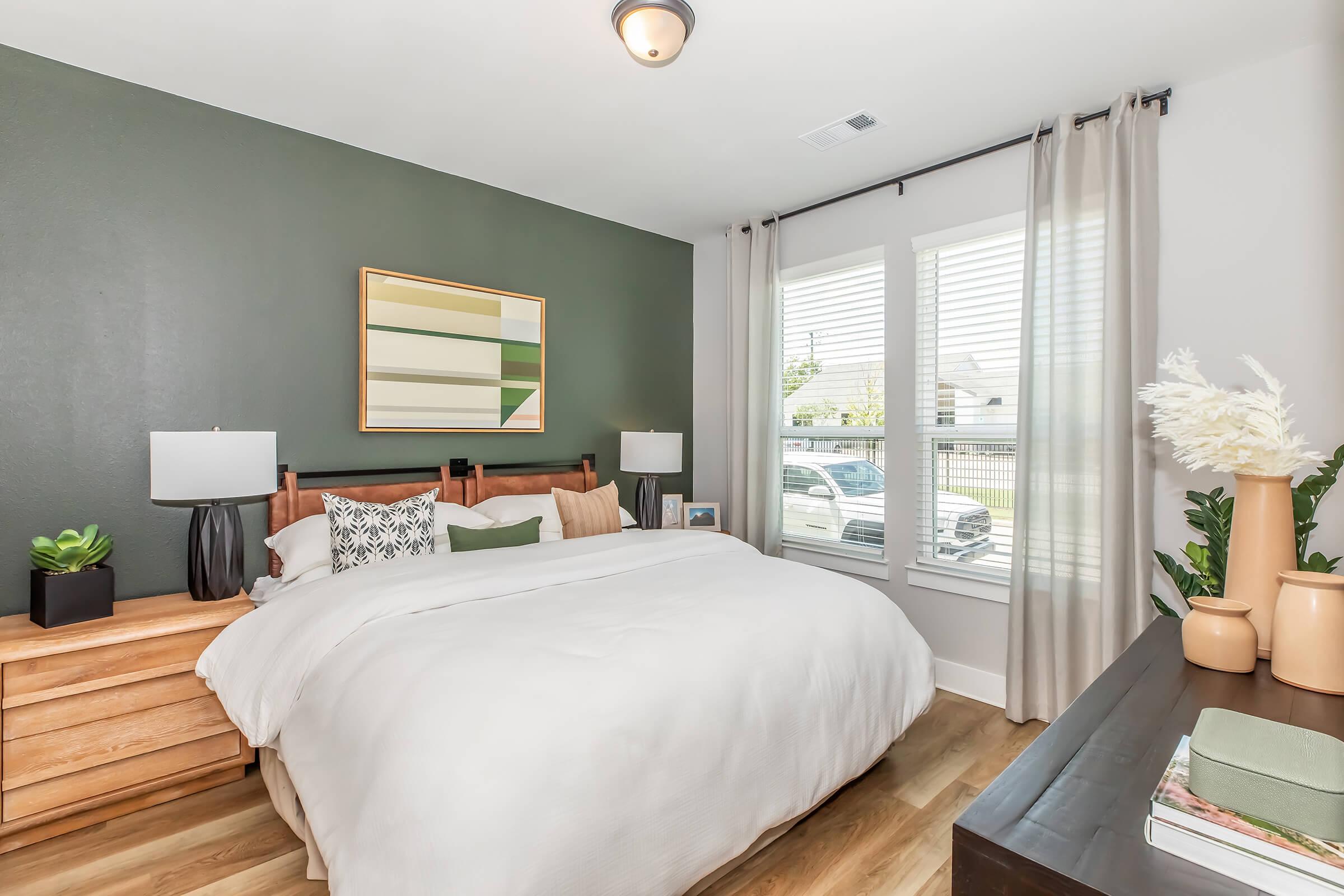 A modern bedroom featuring a wooden bed with white bedding, decorative pillows, and two bedside lamps. The walls are painted a deep green, complemented by a framed abstract art piece. A window with sheer curtains allows natural light in, and a dresser and potted plants add a cozy touch.