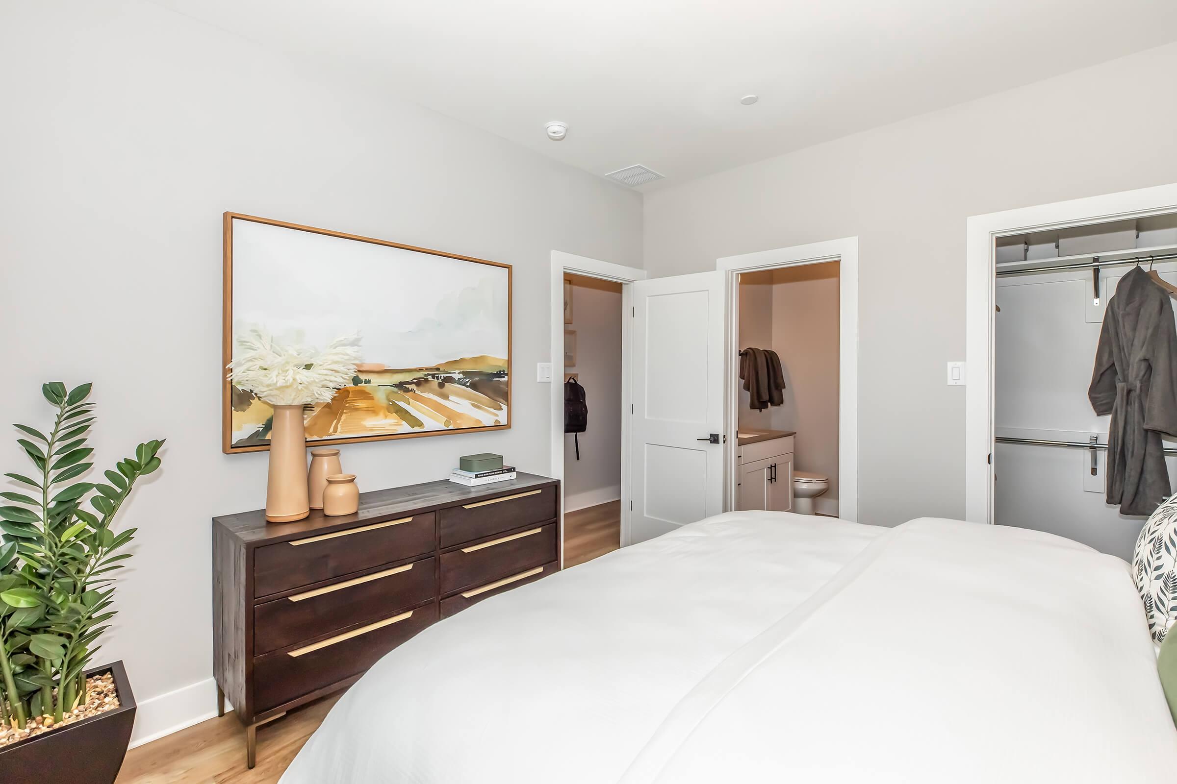 A modern bedroom featuring a large bed with white bedding, a decorative plant in a corner, and a stylish wooden dresser. A framed landscape painting adorns the wall above the dresser. There are two doors; one leads to a closet and the other to a bathroom. The walls are painted a light gray, creating a serene atmosphere.