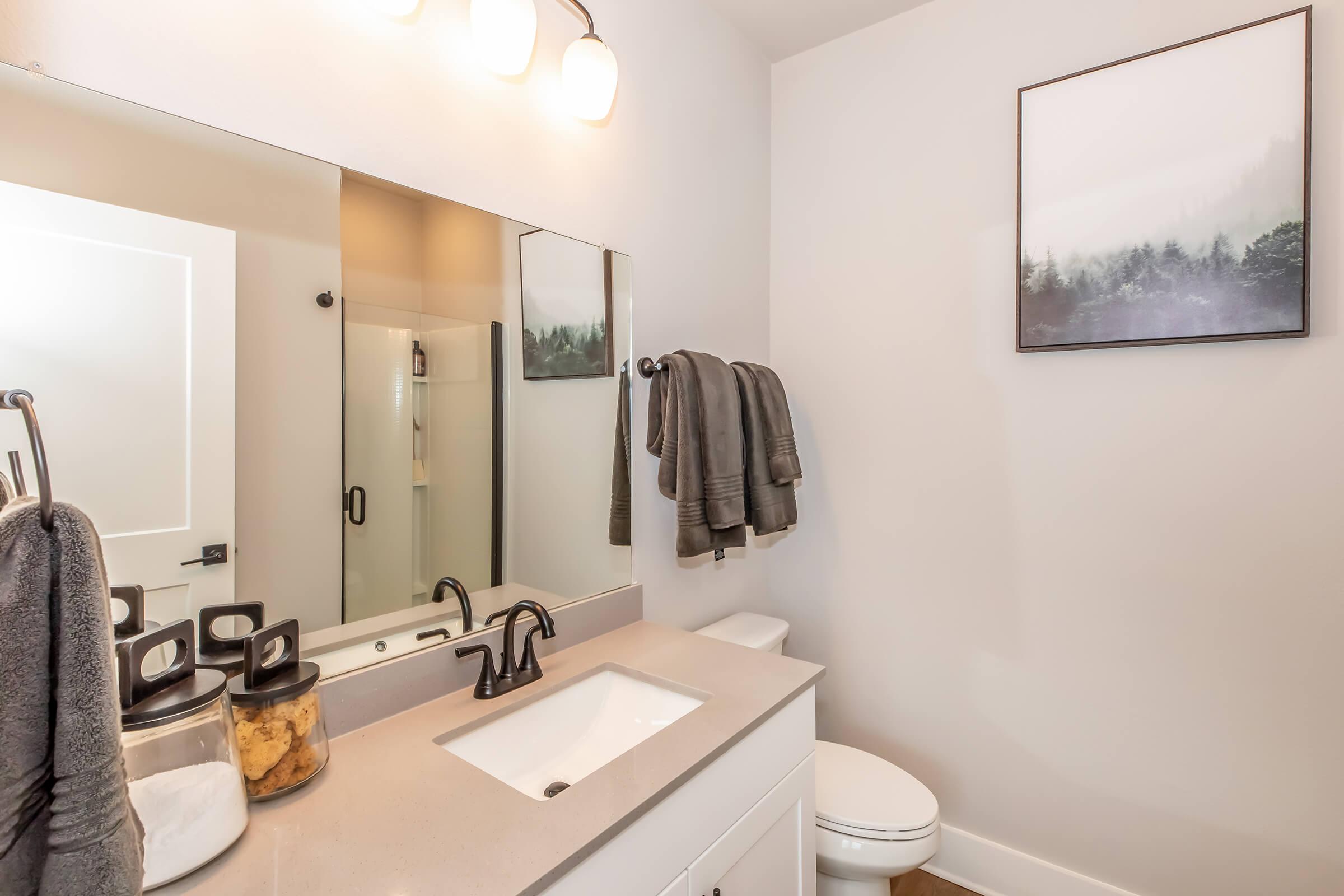 A modern bathroom featuring a white vanity with a rectangular sink, a large mirror, and three light fixtures above. There are neatly hung grey towels, a decorative black-framed landscape print on the wall, and a toilet in the corner. Storage jars with an unclear contents sit on the counter.