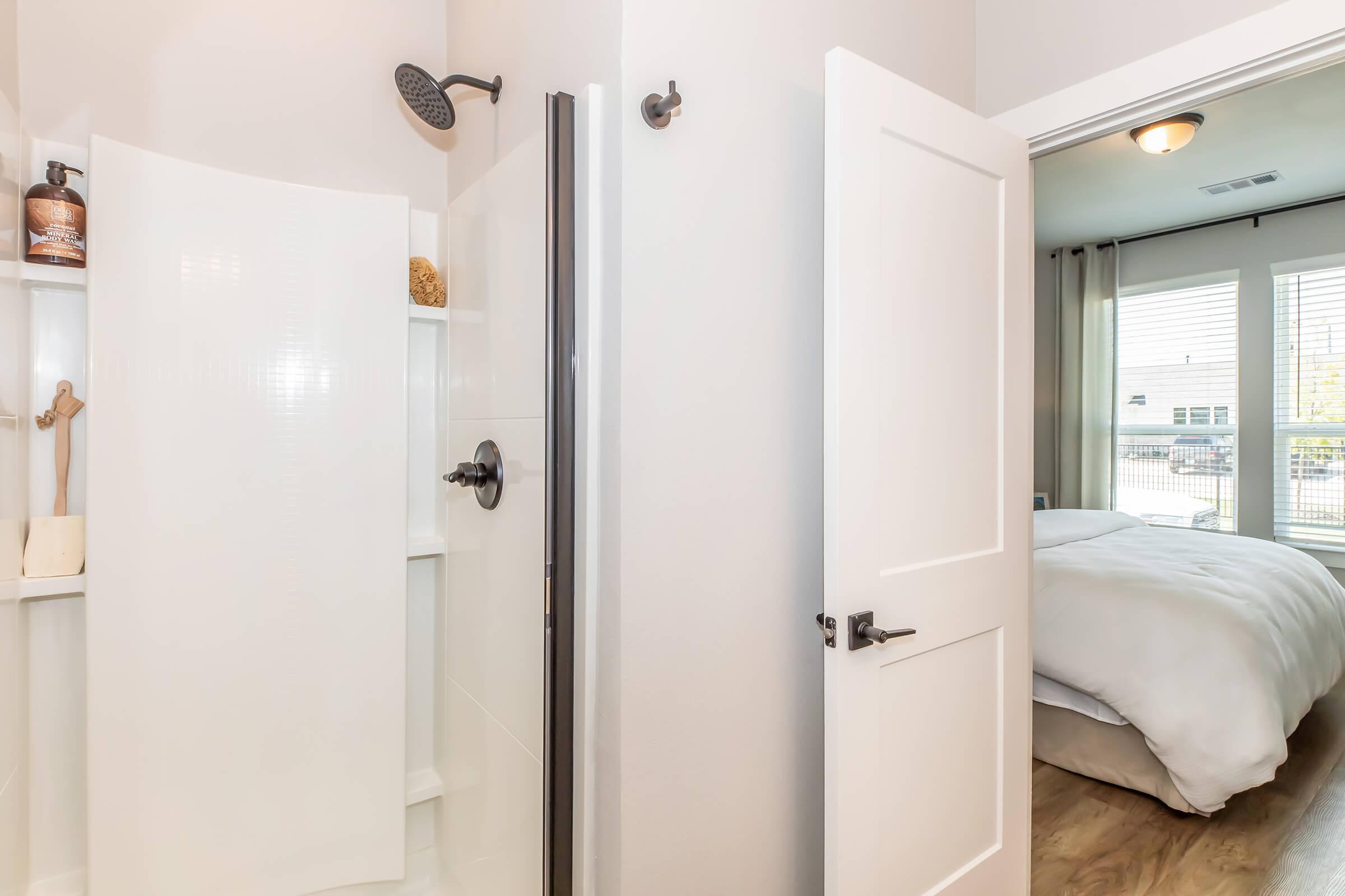 A modern bathroom featuring a glass shower stall with a rain showerhead. The walls are light-colored, and there are shelves with toiletries. In the background, a well-lit bedroom is visible, showing a bed with white bedding and large windows with curtains. The floor is a warm wood tone.